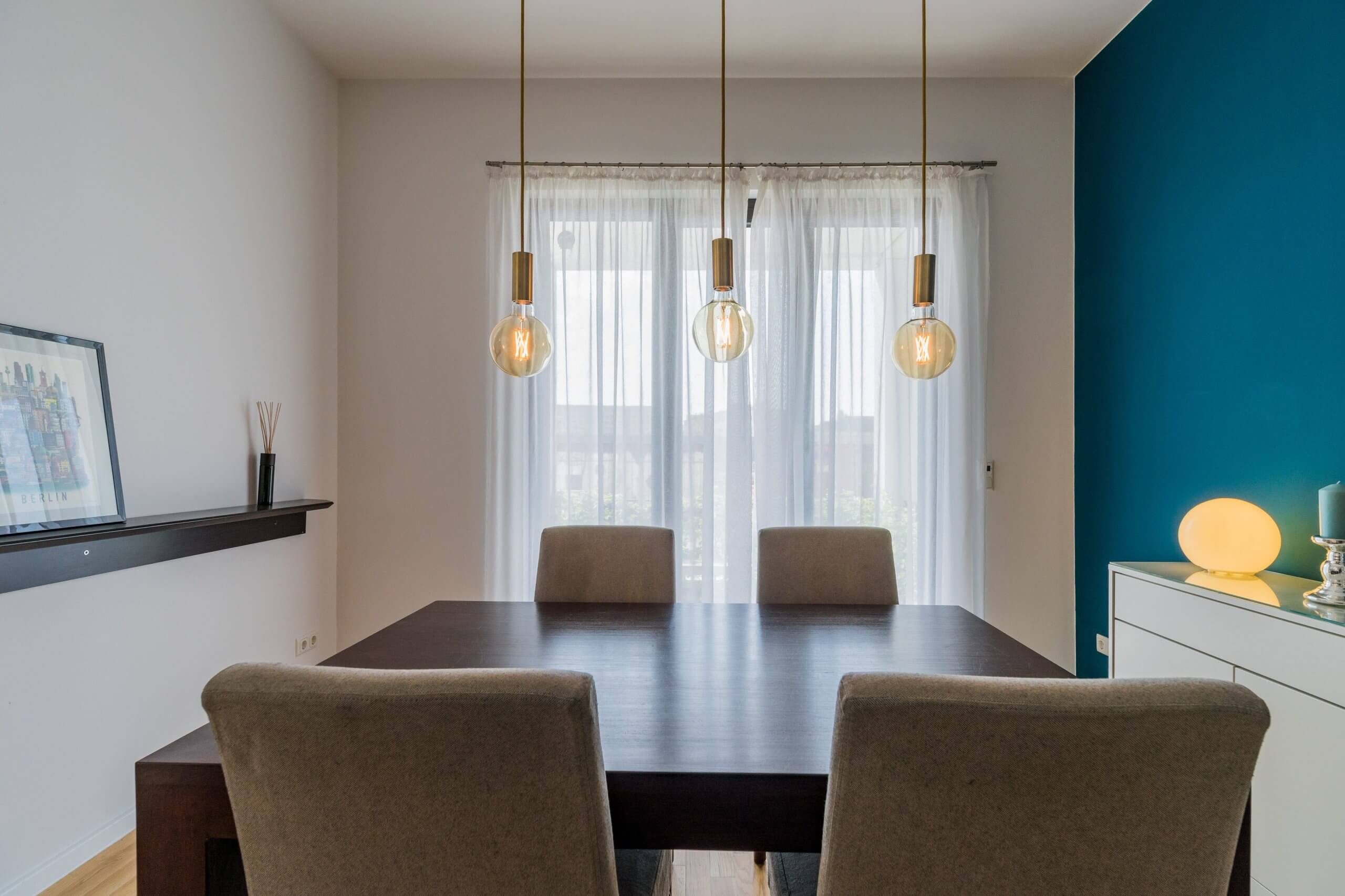 एक अपार्टमेंट के डाइनिंग रूम का सामने से दृश्य, जिसमें एक गहरे रंग की लकड़ी की मेज, कुर्सियाँ और तीन लटकती रोशनी के नीचे फर्श से छत तक फैली एक खिड़की है।