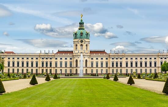 Frontalansicht Barockschloss mit kupfergrüner Kuppel, symmetrischer Gartenanlage und zentralem Springbrunnen