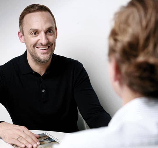 Makler sitzt gegenüber Kunde an Tisch mit Immobilienprospekt in Beratungsgespräch