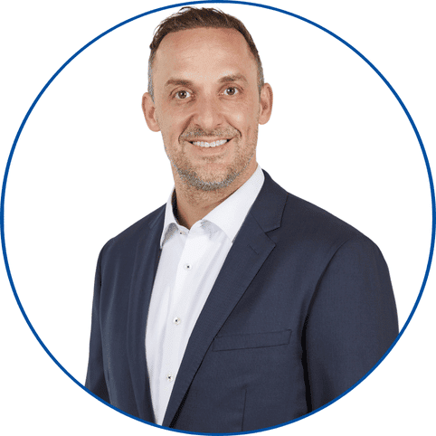 Porträtfoto eines lächelnden Mannes im dunklen Anzug vor weißem Hintergrund in blauumrandetem Kreis