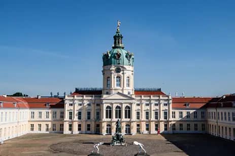 Frontansicht barockes Schloss mit grün patinierter Kuppel und symmetrischen Flügelbauten vor klarem Himmel