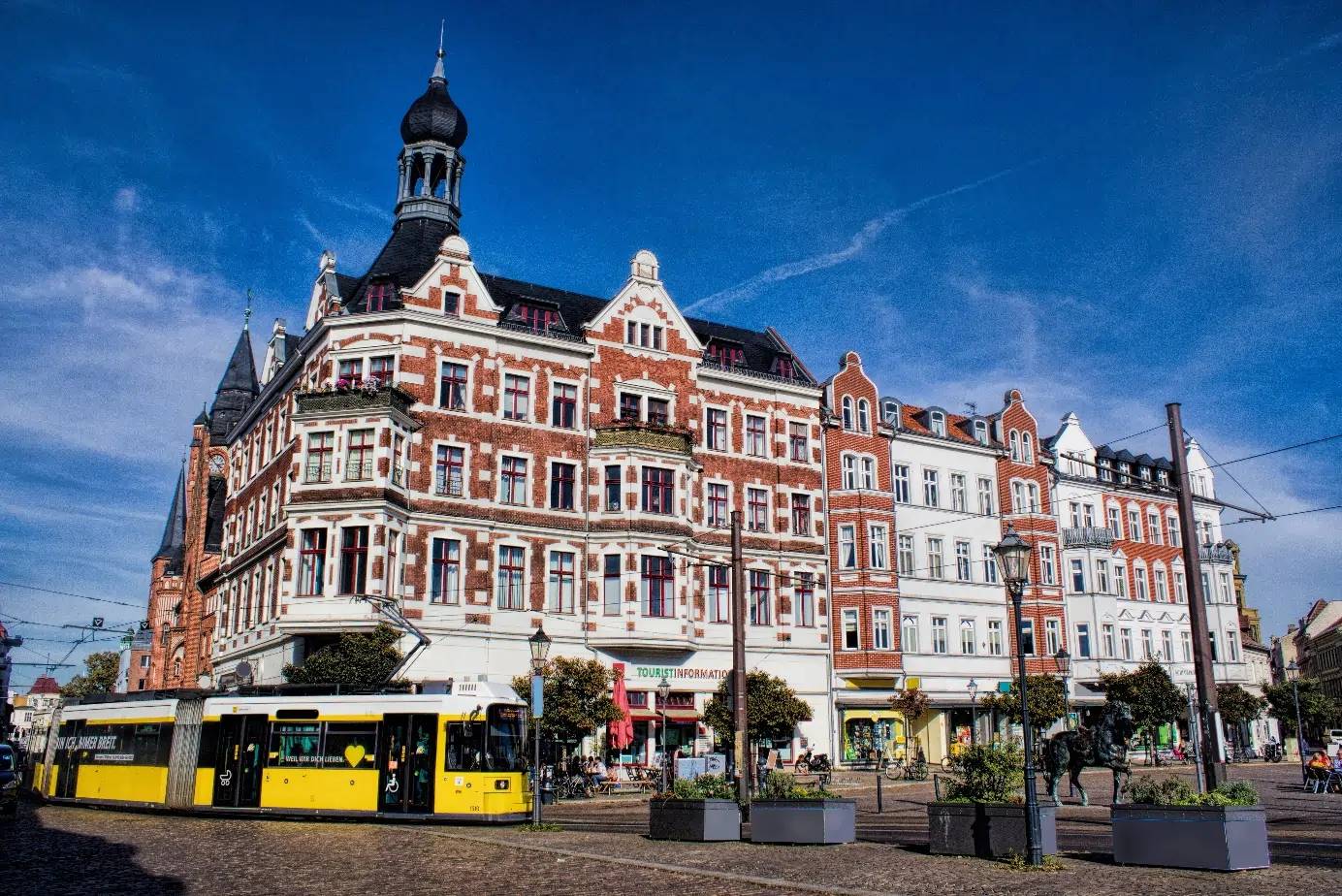 Zabytkowy budynek w słoneczny dzień, żółty tramwaj przejeżdża obok, błękitne niebo w tle. Atmosfera miejskiego życia.