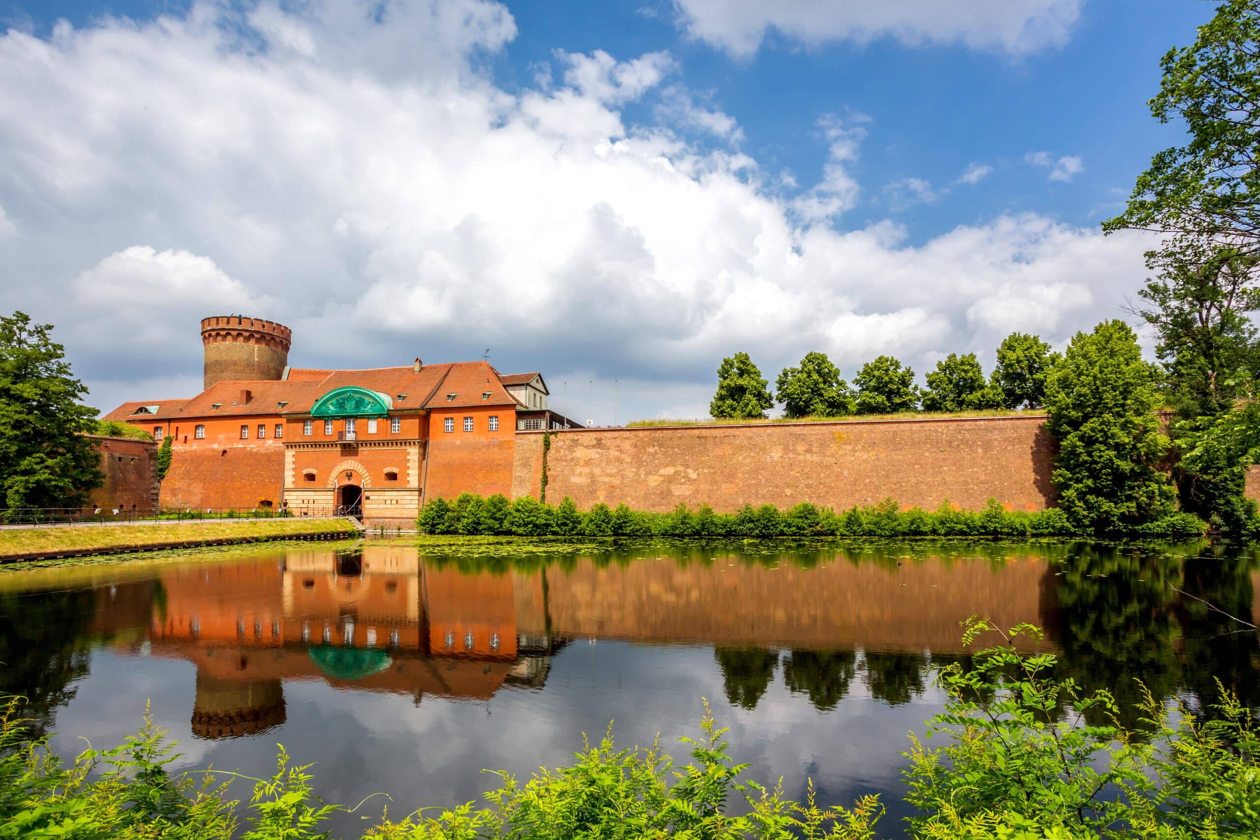 Vorderansicht einer historischen Backsteinfeste mit Rundturm und Wassergraben im Spiegelbild