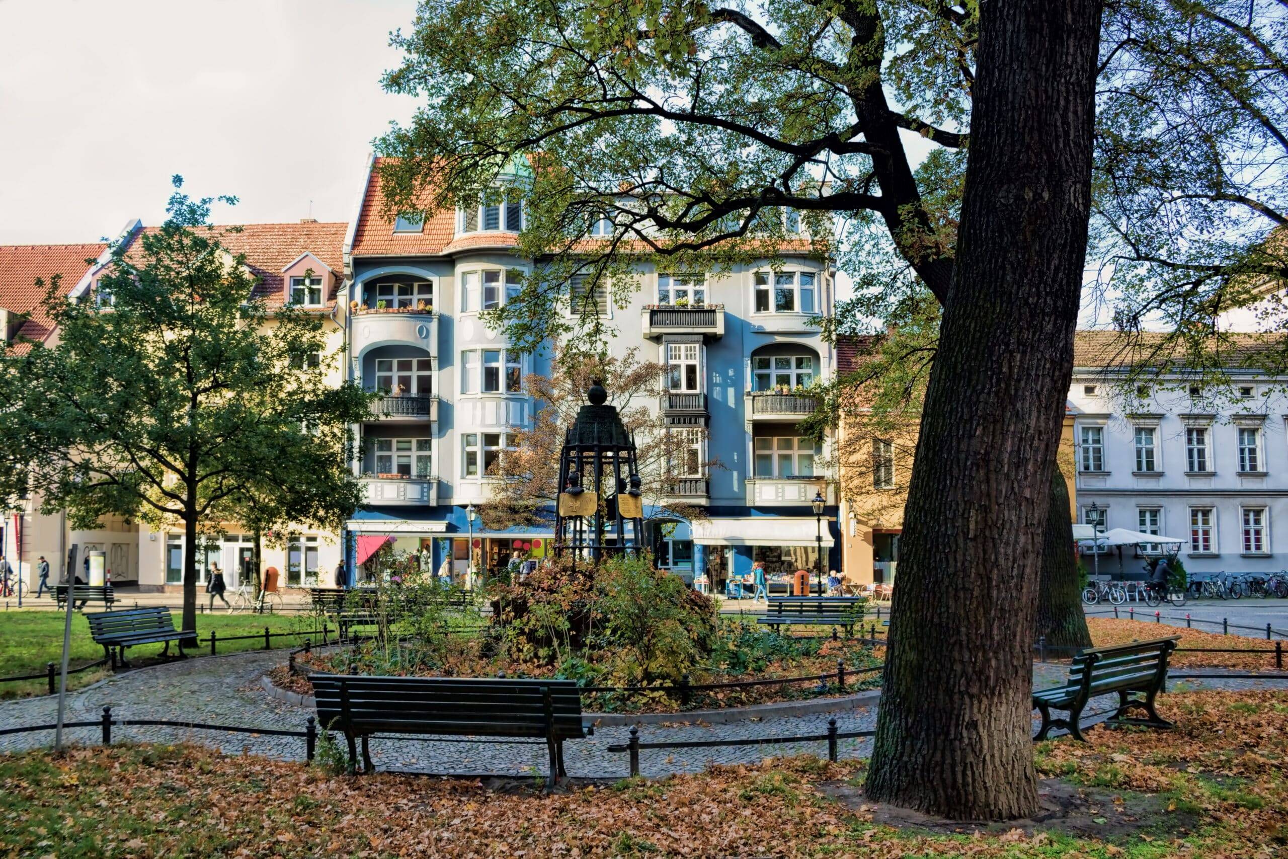 kleiner Park mit Bänken und ornamentalem Brunnen im Vordergrund vor pastellfarbenen Altbauhäusern