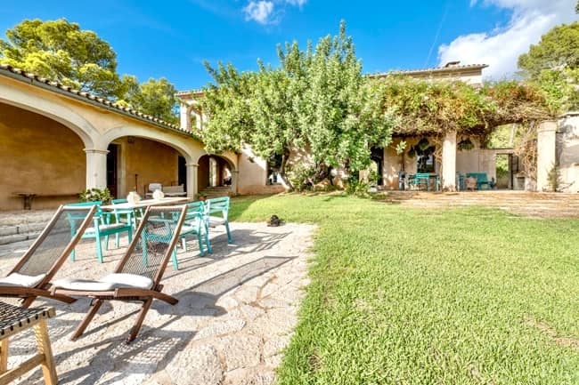 Blick von Steinterrasse auf mediterrane Landvilla mit Säulenbögen, Weinranken und Gartenmöbel auf Rasen