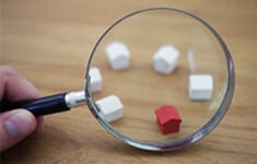 Große Lupe über rotem Miniaturhaus umgeben von weißen Modellhäusern auf Holzuntergrund