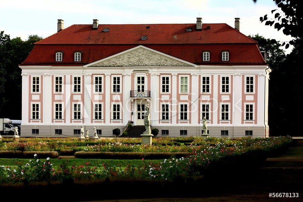 Frontansicht eines barocken Herrenhauses mit rosaweißer Fassade rotem Ziegeldach Balkon und klassizierendem Rosengarten