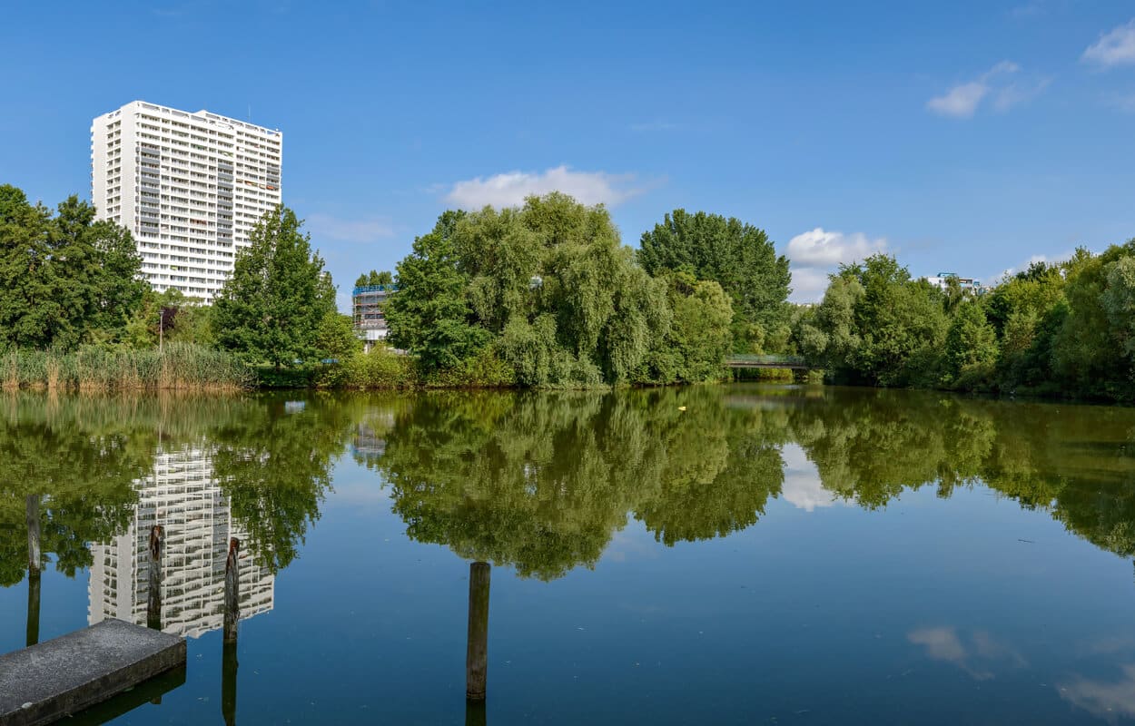 Blick auf weißes Wohnhochhaus am Seeufer mit Spiegelung im Wasser und umgebendem Baumbestand