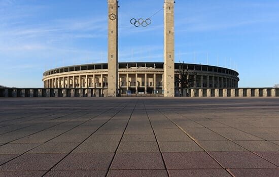 Vooraanzicht van een ovaal stadion met twee torenkolommen en hangende Olympische ringen
