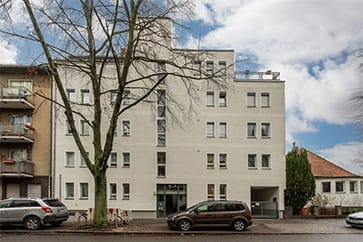 Frontansicht Mehrfamilienhaus mit weißer Fassade und Flachdach aus Straßenperspektive mit Straßenbaum und parkenden Autos