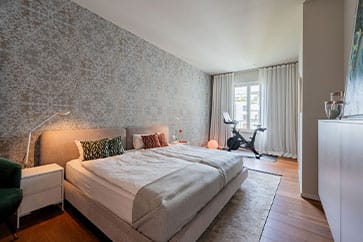 Innenansicht eines modernen Schlafzimmers mit Doppelbett, gemusterter Wand, bodentiefen Fenstern und Fitnessbike auf Parkett