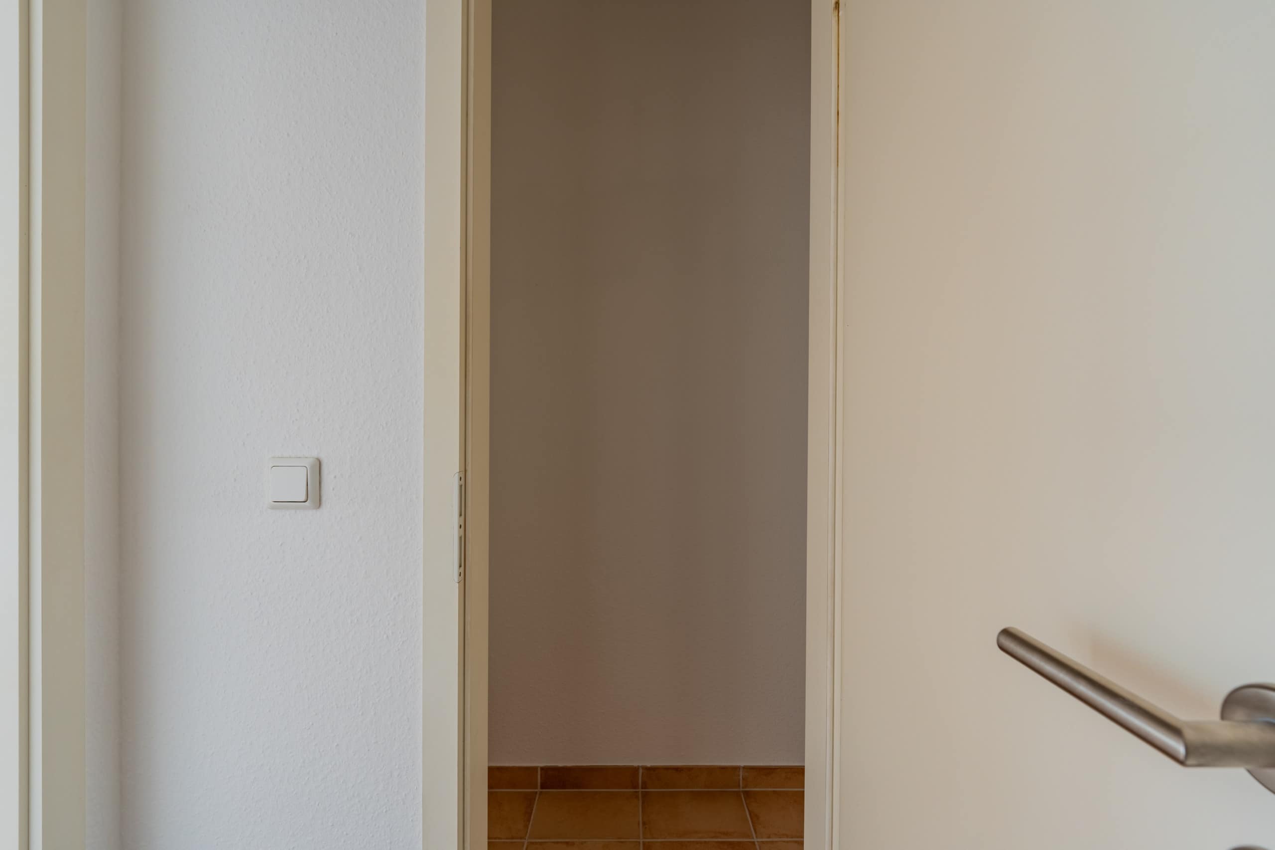 Detailaufnahme geöffneter weißer Innentür mit Edelstahlgriff und Lichtschalter an verputzter Wand über Fliesenboden