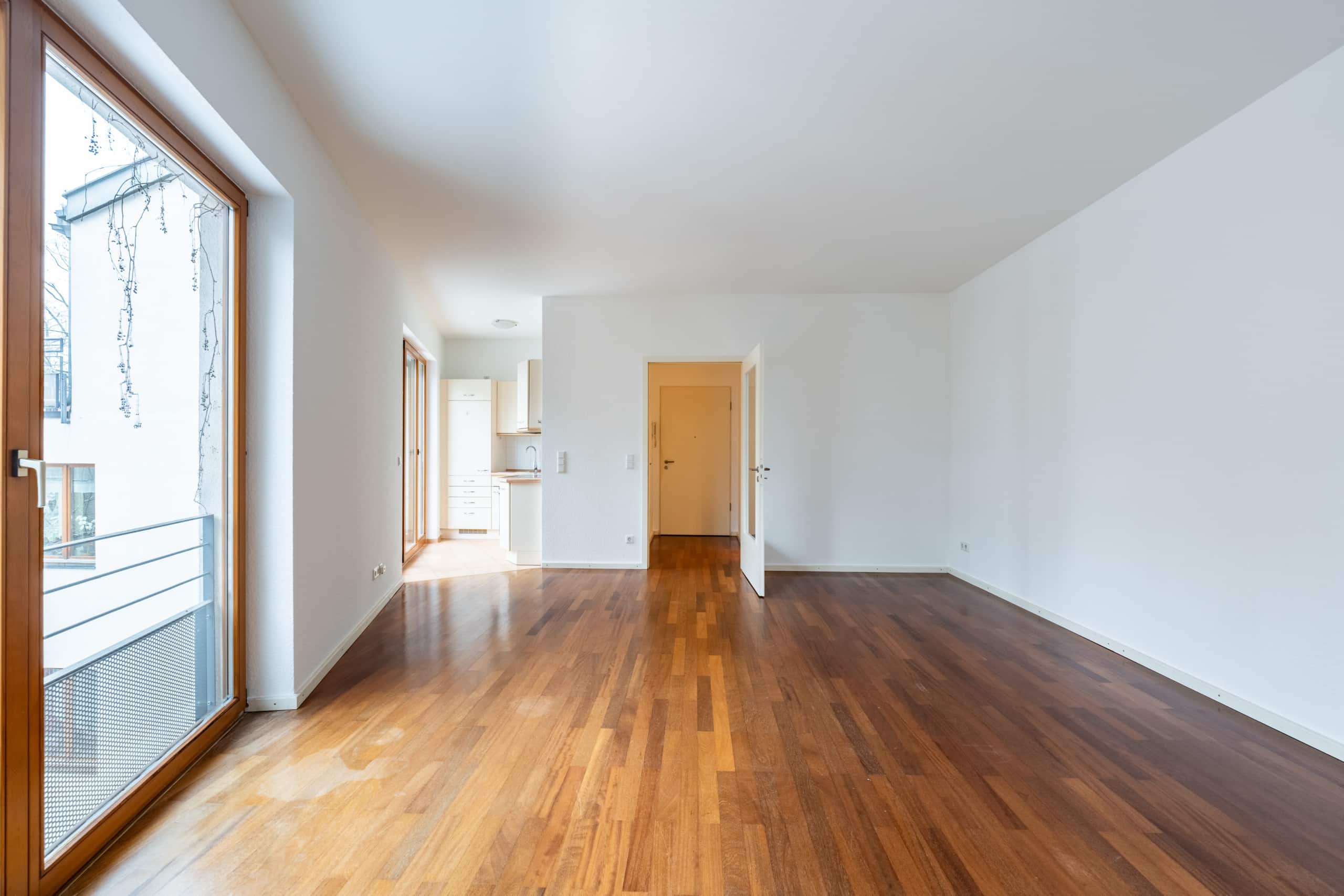 Blick ins helle Wohn­zimmer einer Neubauwohnung mit Parkettboden bodentiefen Fenstern Balkon und offener Einbauküche