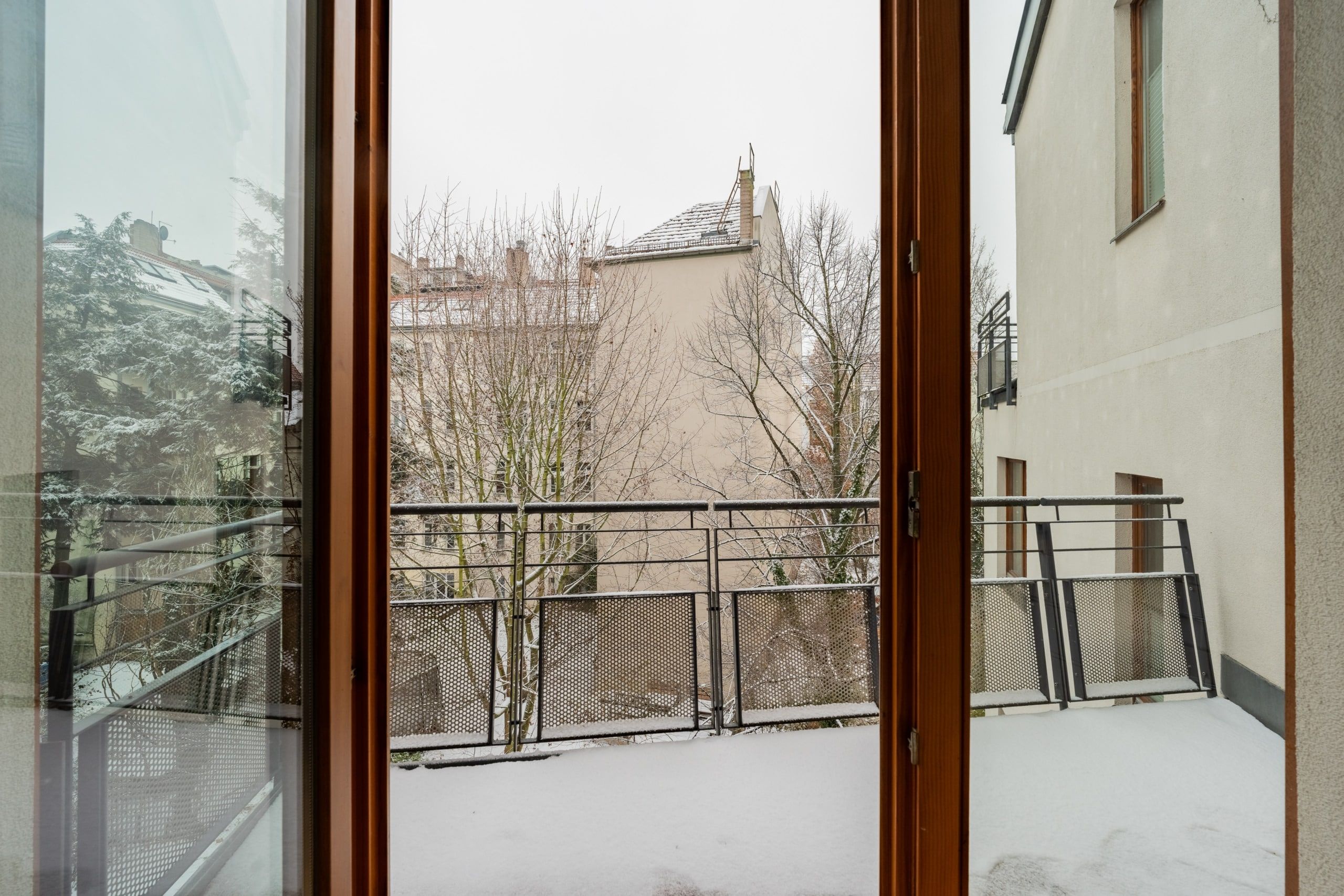 Blick durch geöffnete Terrassentür auf schneebedeckten Balkon mit Metallgeländer und Nachbargebäude