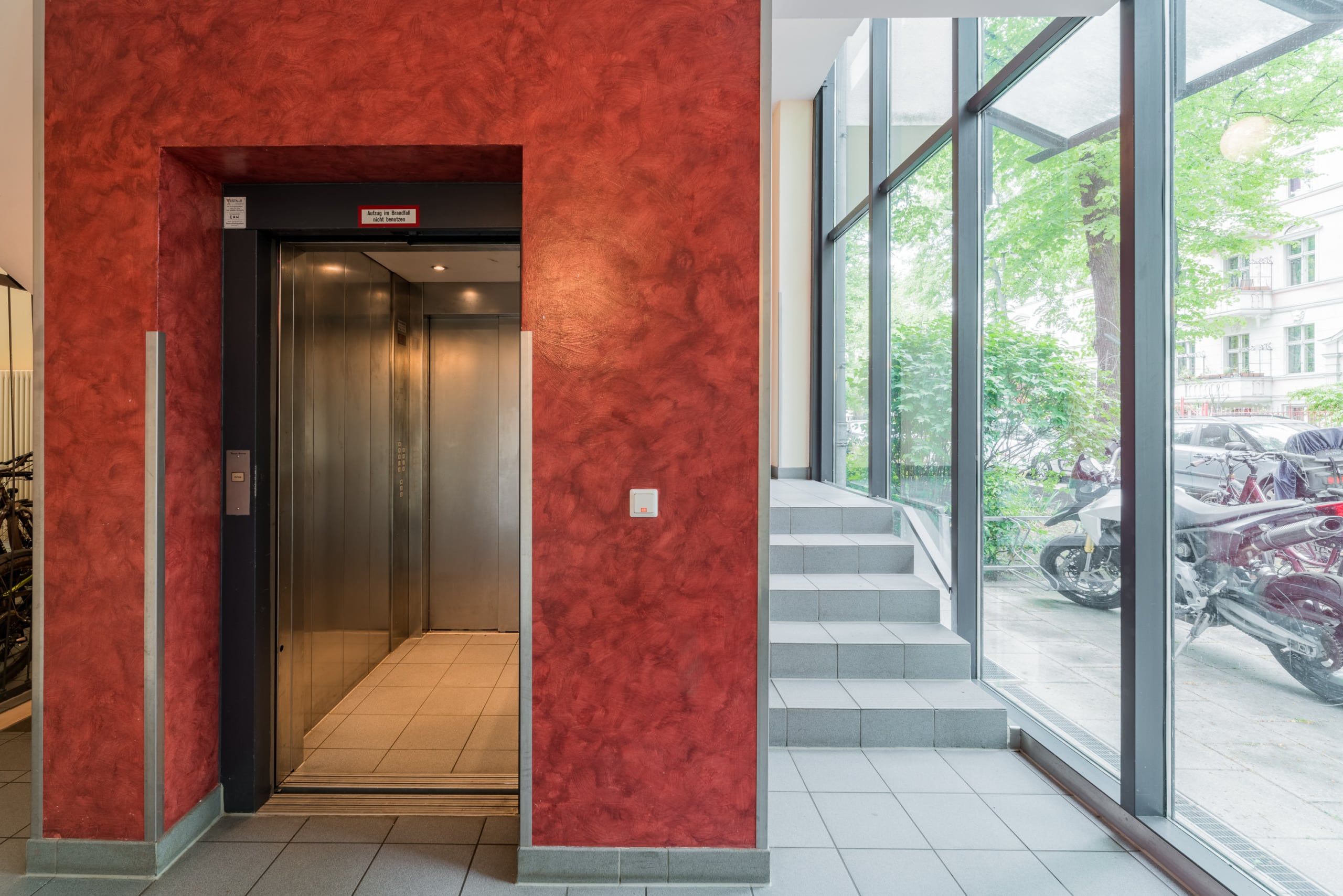 Lobby eines Mehrfamilienhauses mit offenem Aufzug, roter Akzentwand, Treppe und bodentiefer Glasfront zum Außenbereich