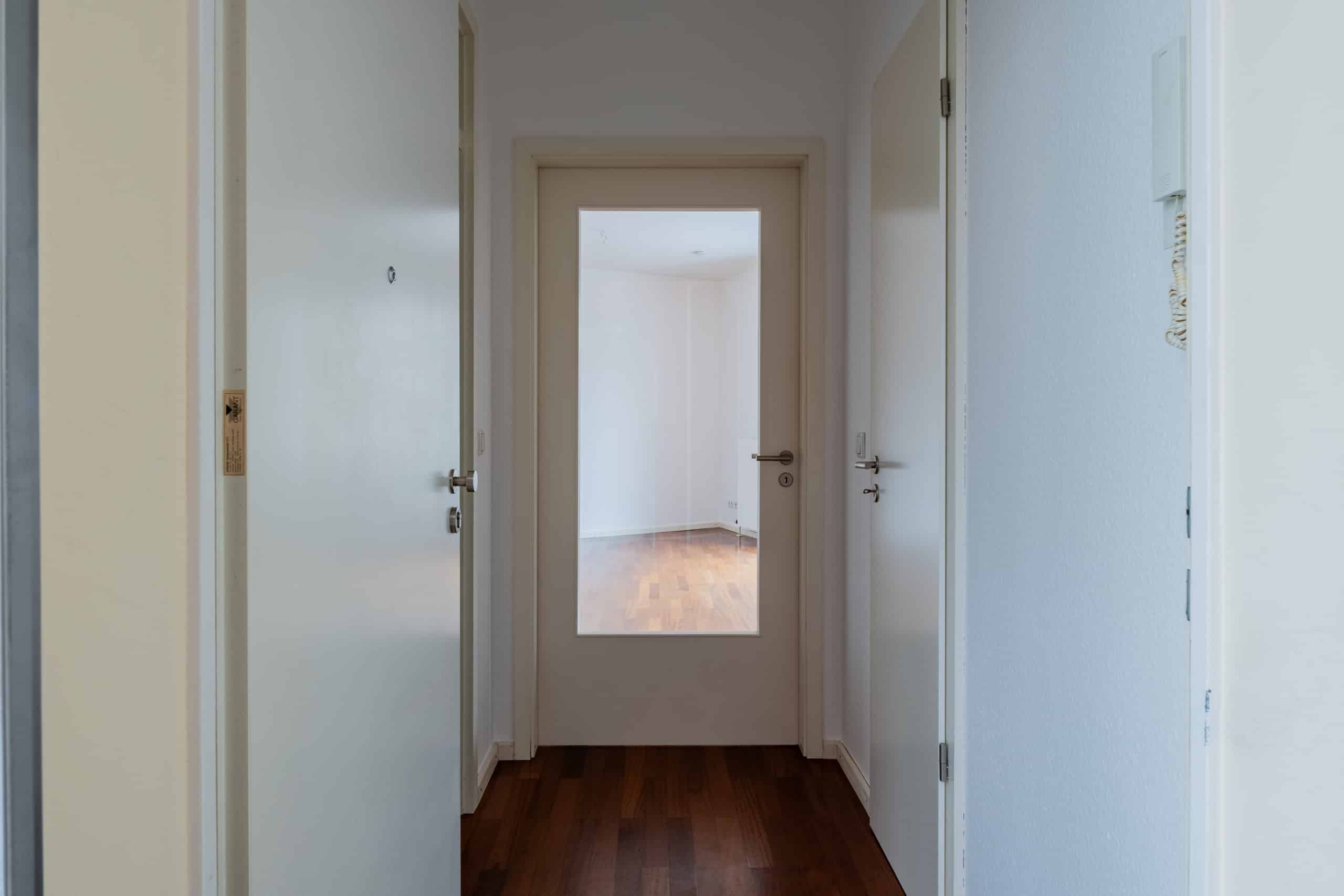 Innenansicht eines Wohnungsflurs mit weißer Haustür links, Glastür geradeaus und dunklem Parkettboden