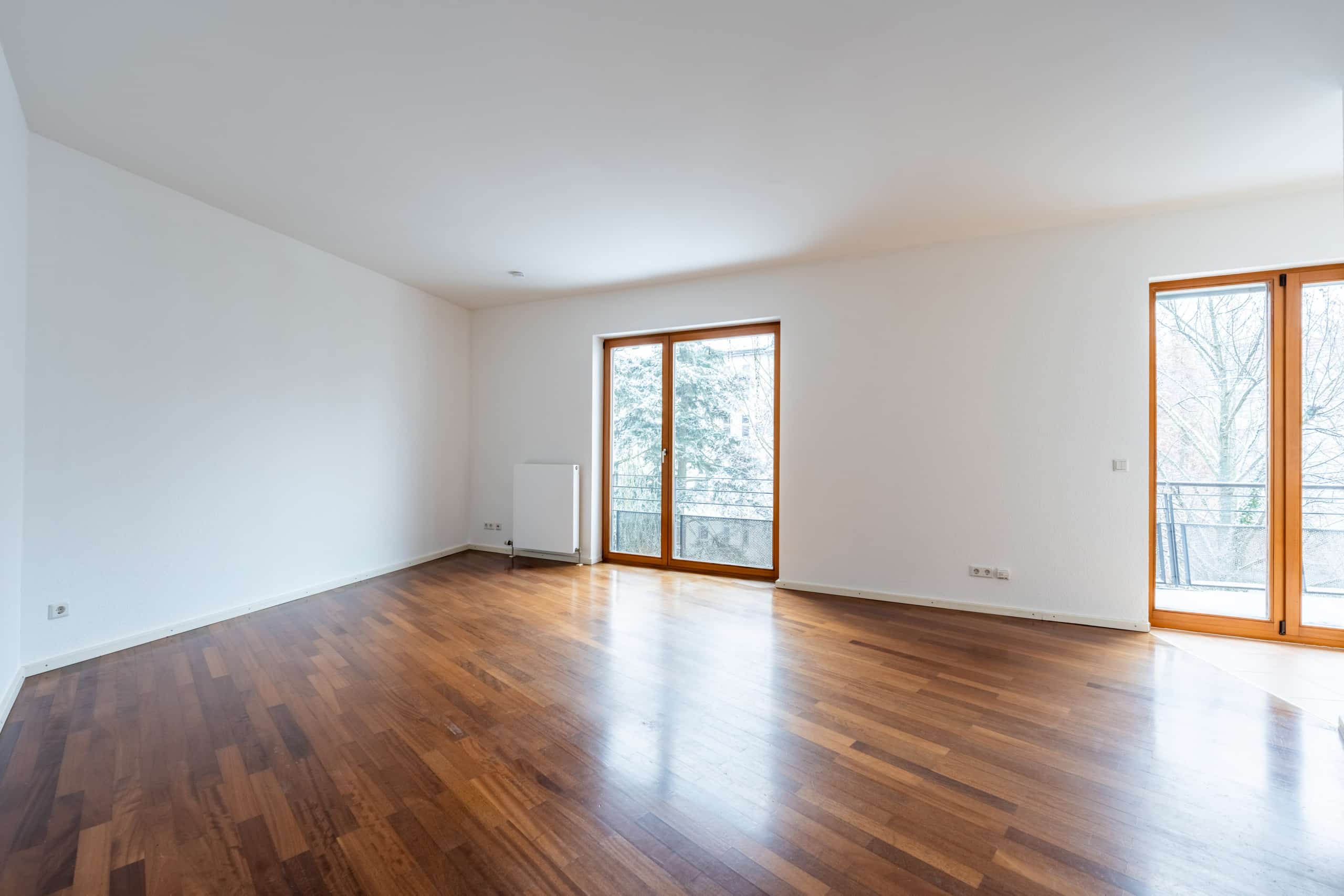 großer leerer Wohnraum mit dunklem Parkettboden und zwei bodentiefen Fenstertüren zu kleinem Balkon