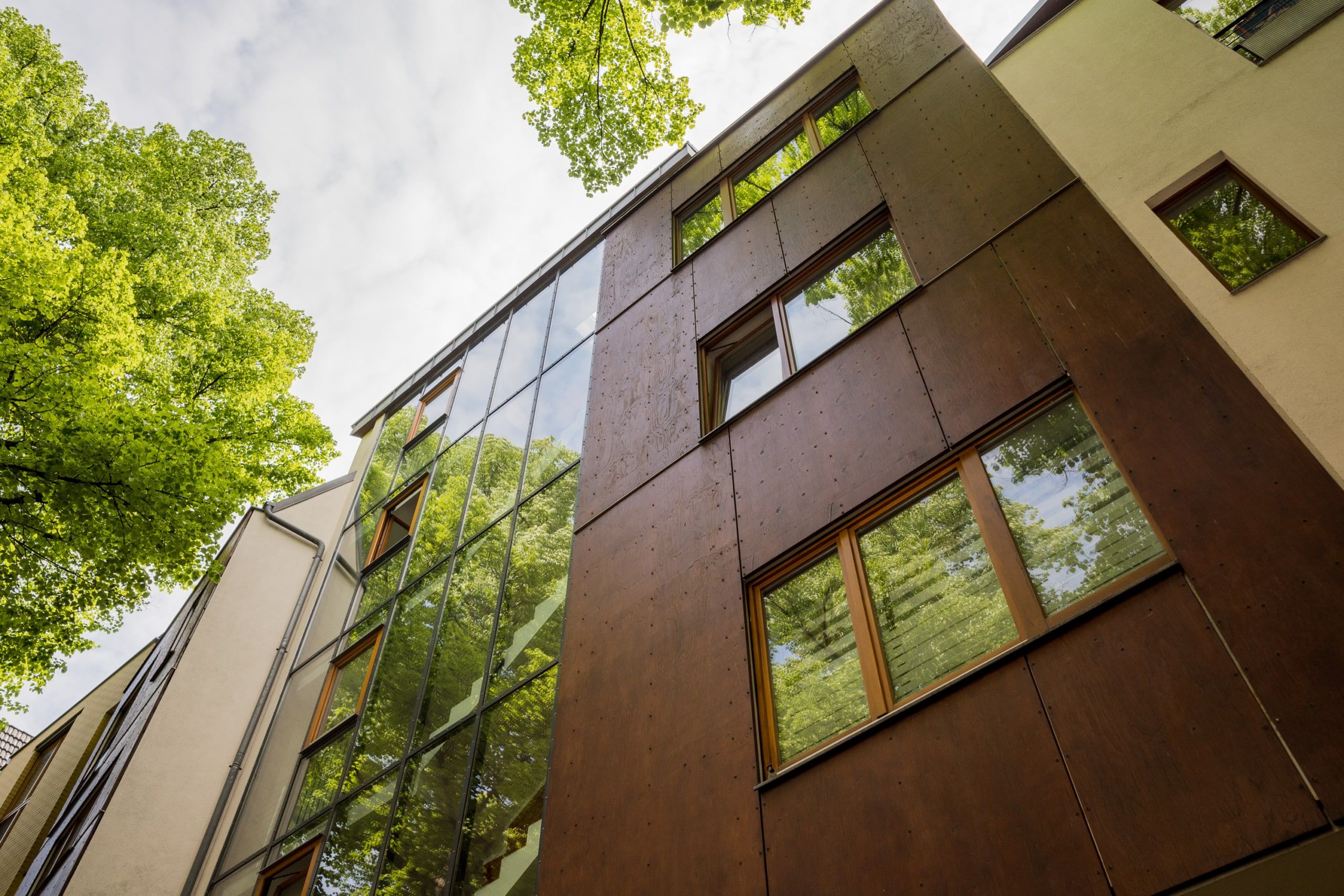 Moderne mehrgeschossige Wohnhausfassade in Froschperspektive mit Glasfront, dunkelbraunen Holzplatten und Baumsilhouetten