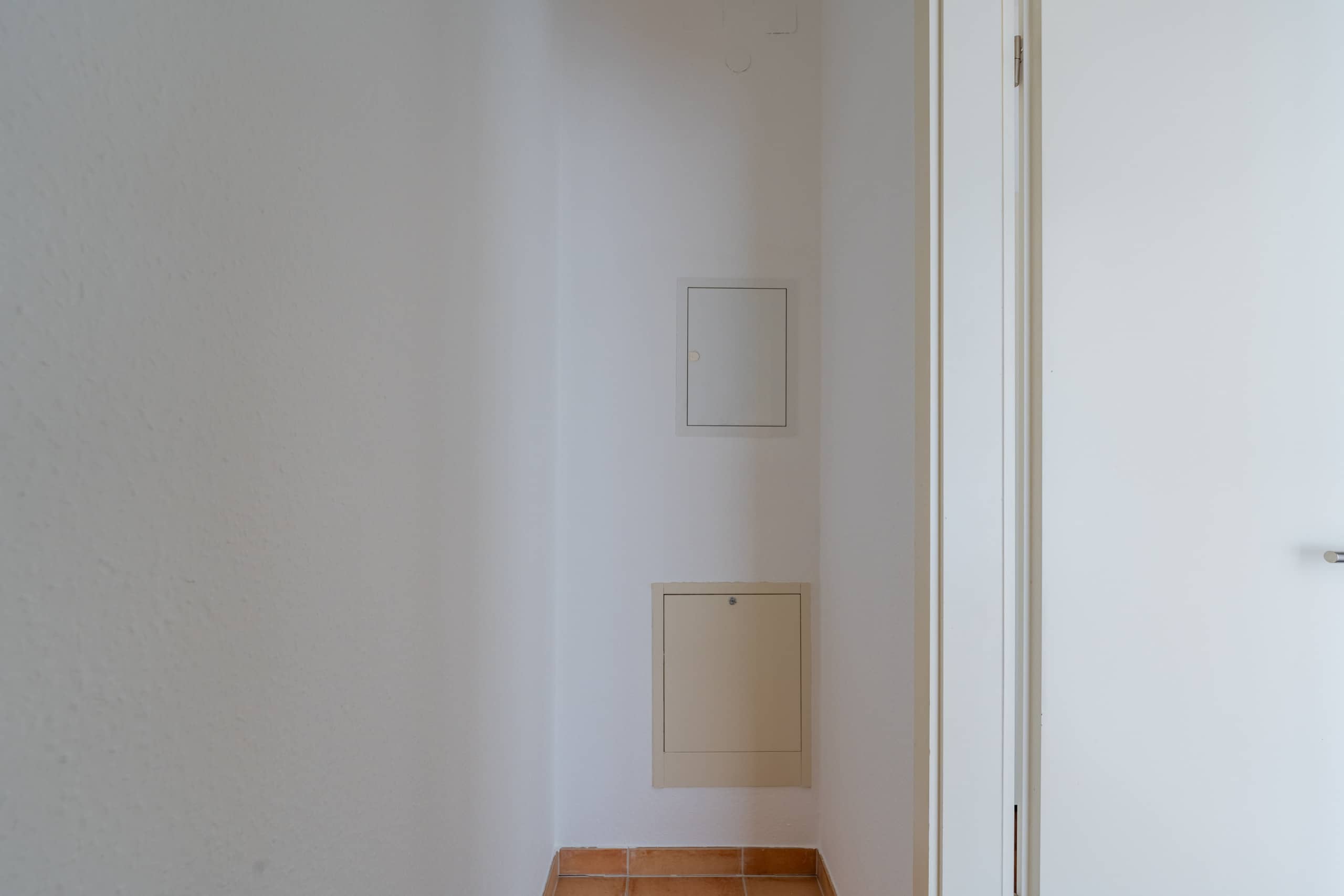 schmaler Hausflur mit zwei Zugangspaneelen an Wand mit offenem Türrahmen rechts und terrakottafarbenen Bodenfliesen