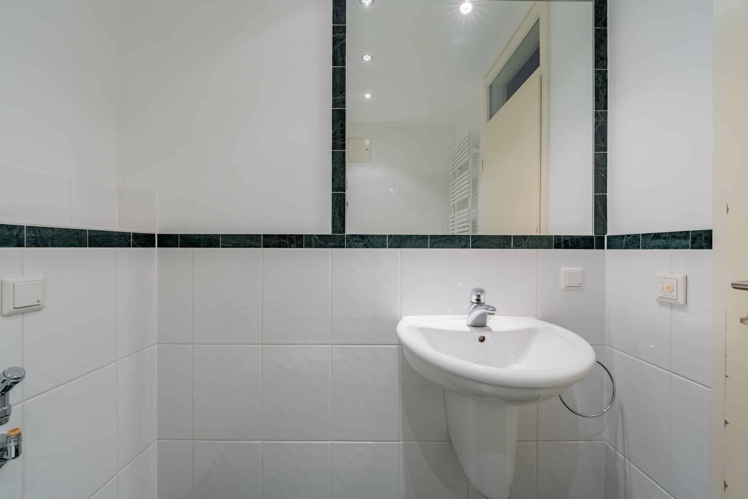 Innenperspektive Badezimmer mit weißem Wandwaschbecken unter großem Spiegel und dunkelgrüner Fliesenbordüre