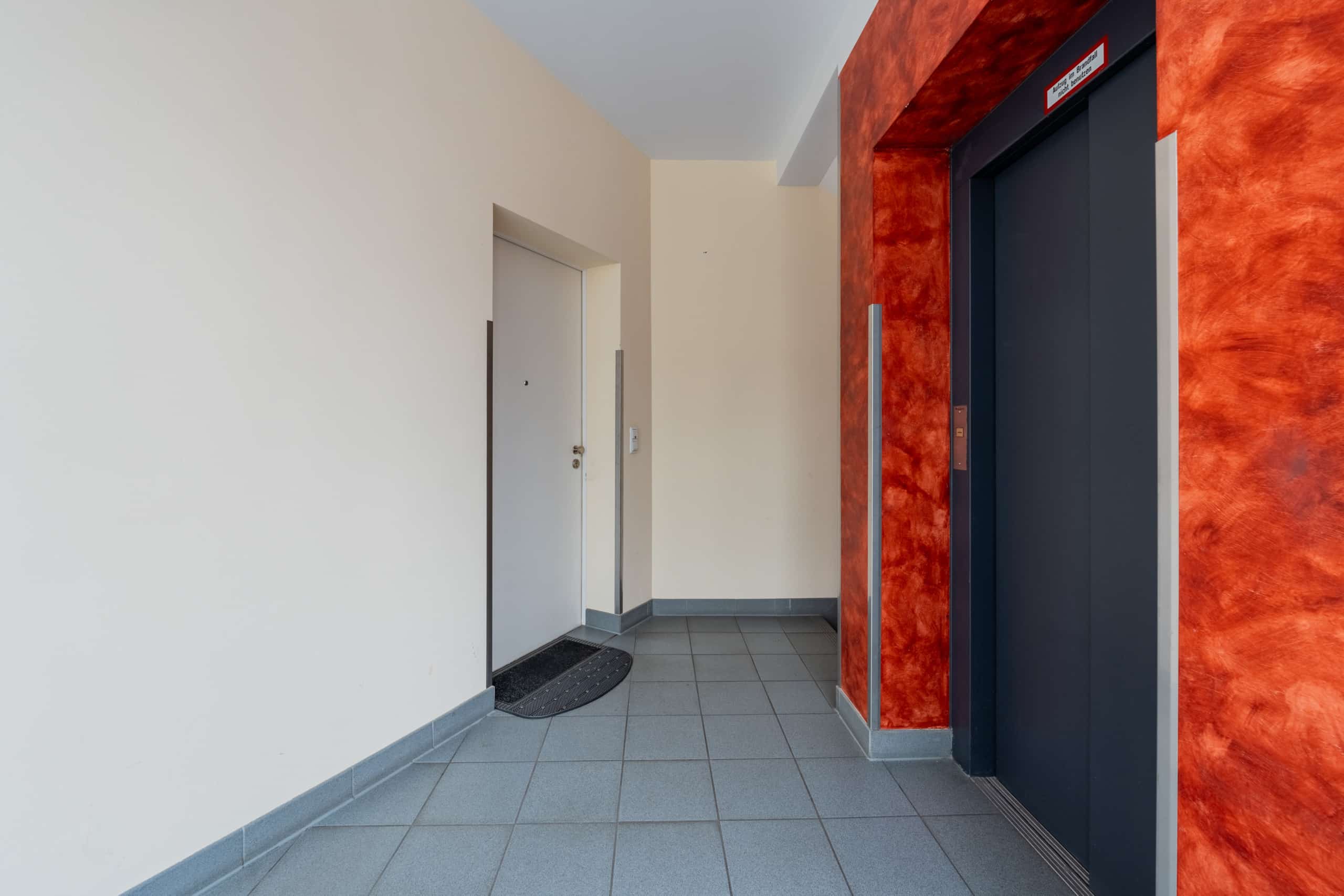 Frontale Innenansicht Hausflur mit grauen Bodenfliesen, hellen Wänden, roter Aufzugsnische und weißer Wohnungstür
