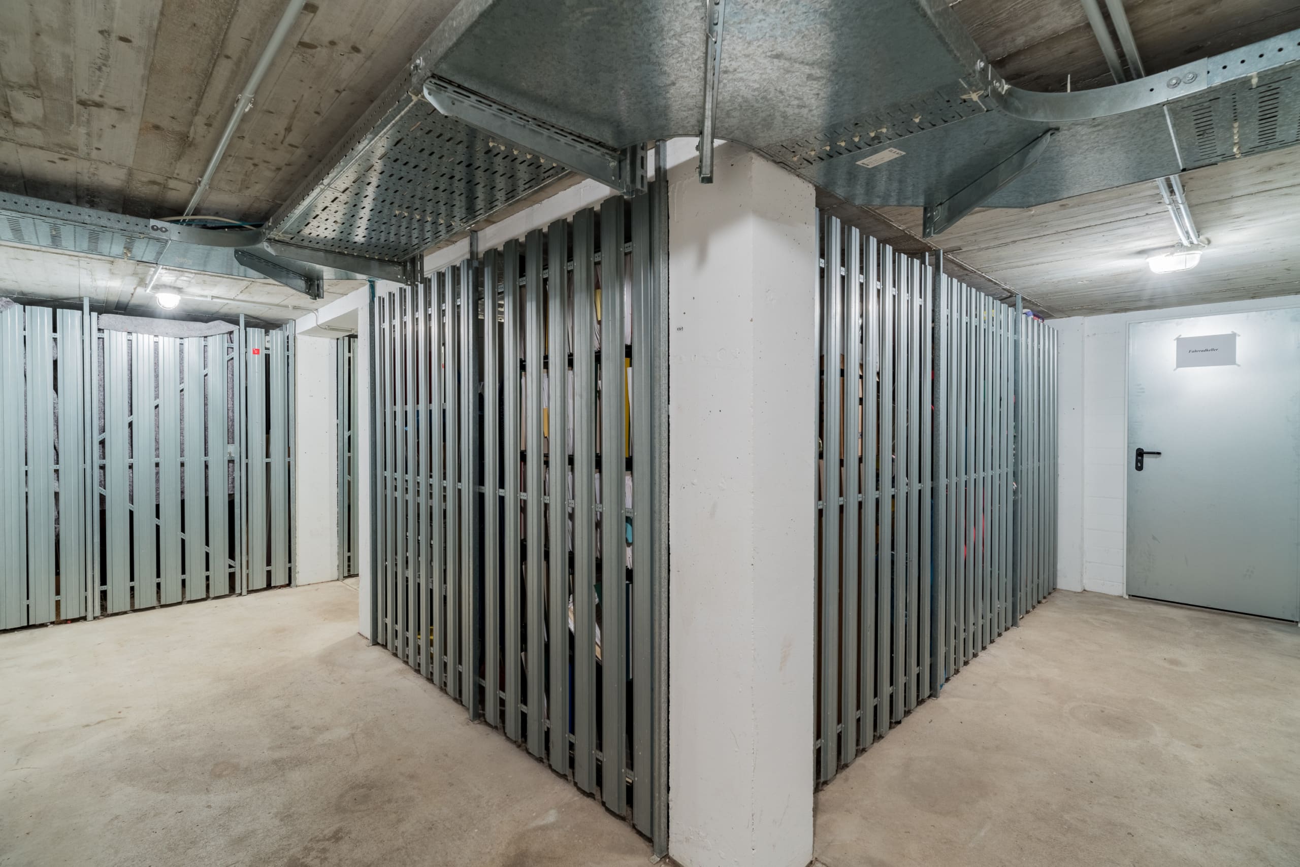 Blick in den Kellerflur eines Mehrfamilienhauses mit gitterartigen Abstellkäfigen und Tür zum Fahrradkeller