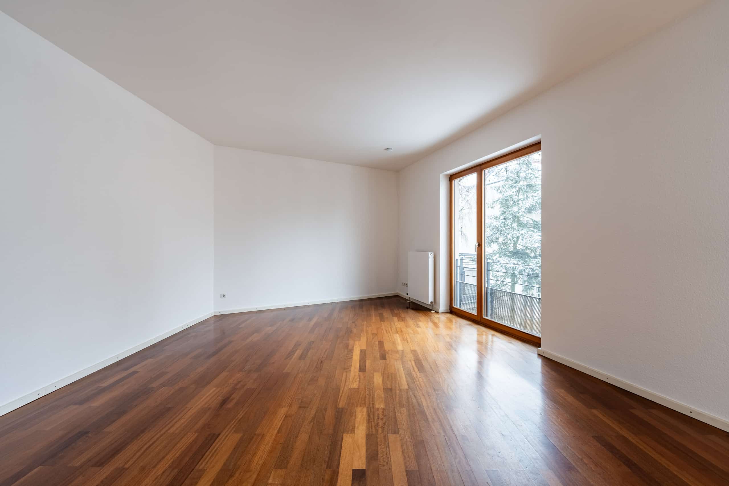 Innenansicht eines leerstehenden Wohnzimmers mit Parkettboden und bodentiefer Fensterfront zum Balkon