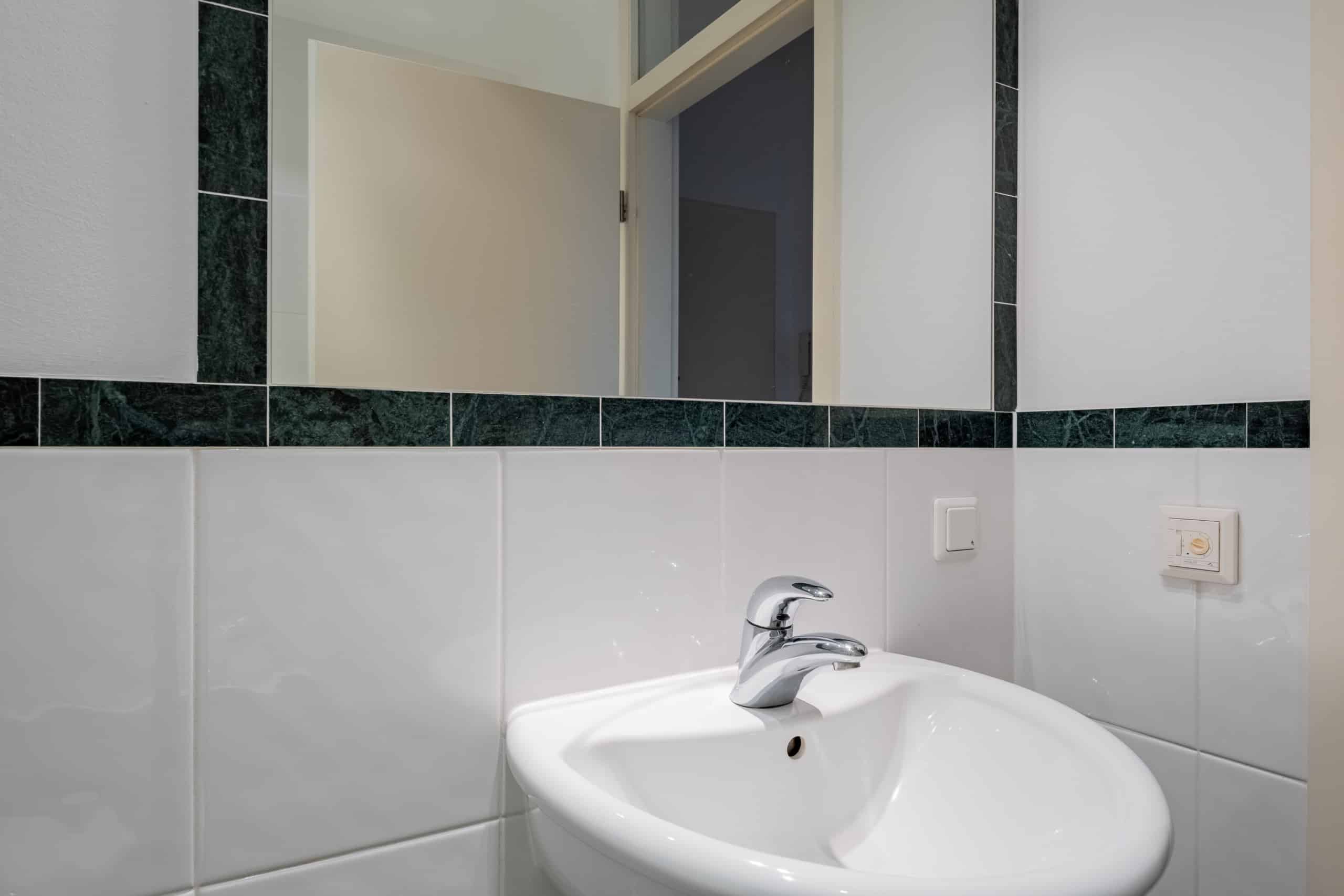 Detailansicht Badezimmer mit weißem Waschbecken, Chromarmatur und grünem Marmorfliesenband unter Spiegel