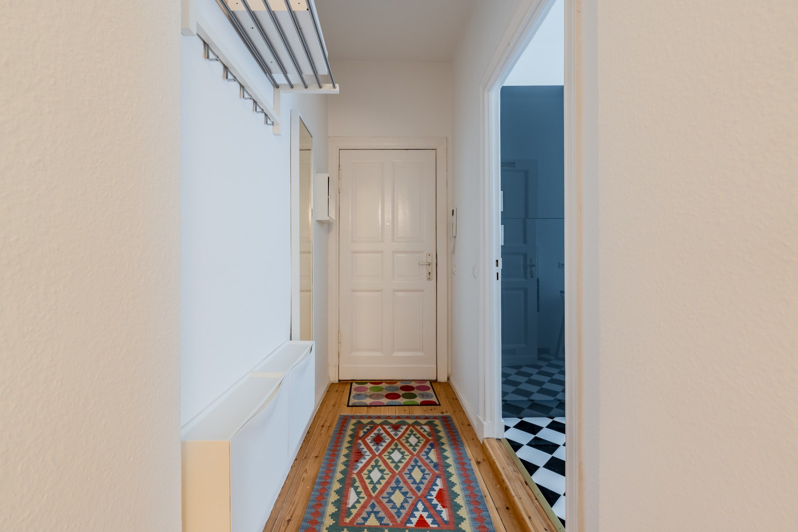 Apartmentflur mit Blick zur weißen Eingangstür Dielenboden bunter Läufer Wandgarderobe und Spiegel