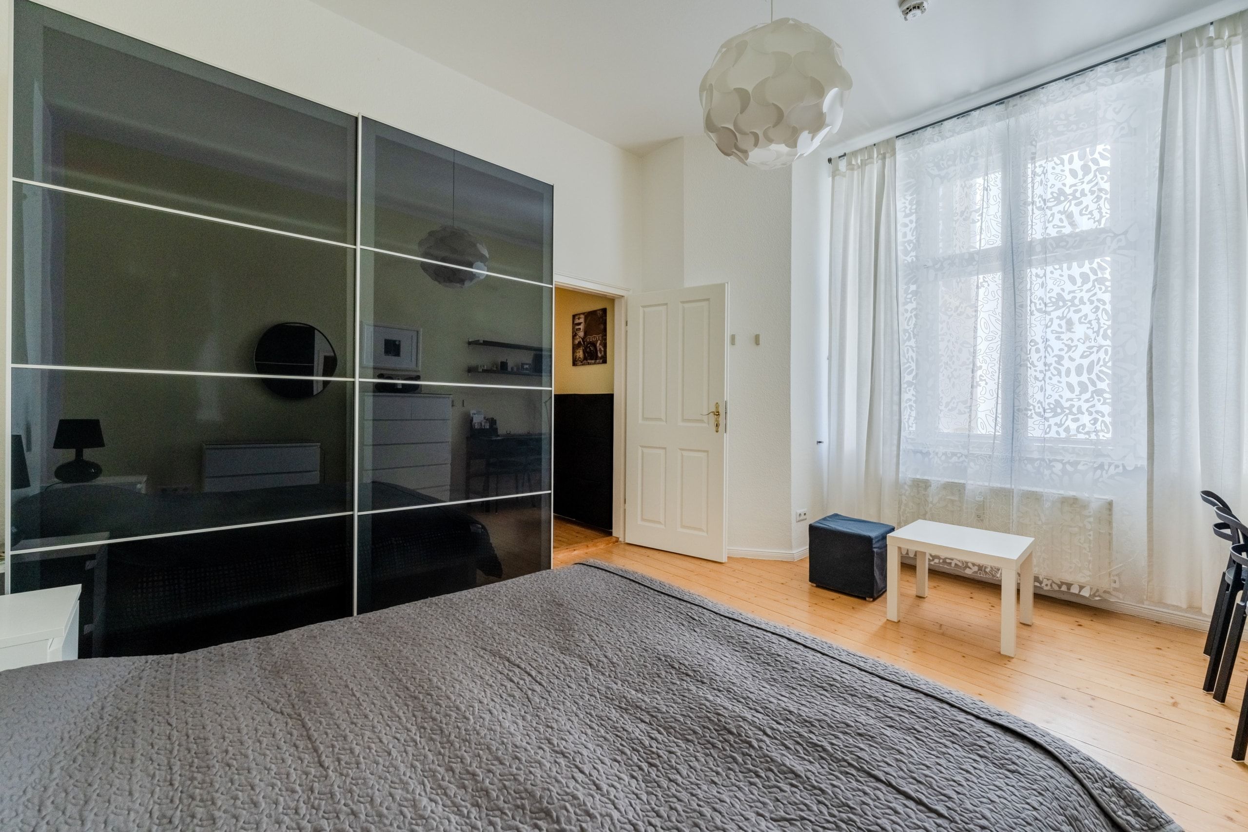 Blick ins Schlafzimmer mit Bett, spiegelndem Schiebetürenschrank, Holzdielen und Fenster mit Spitzenvorhängen