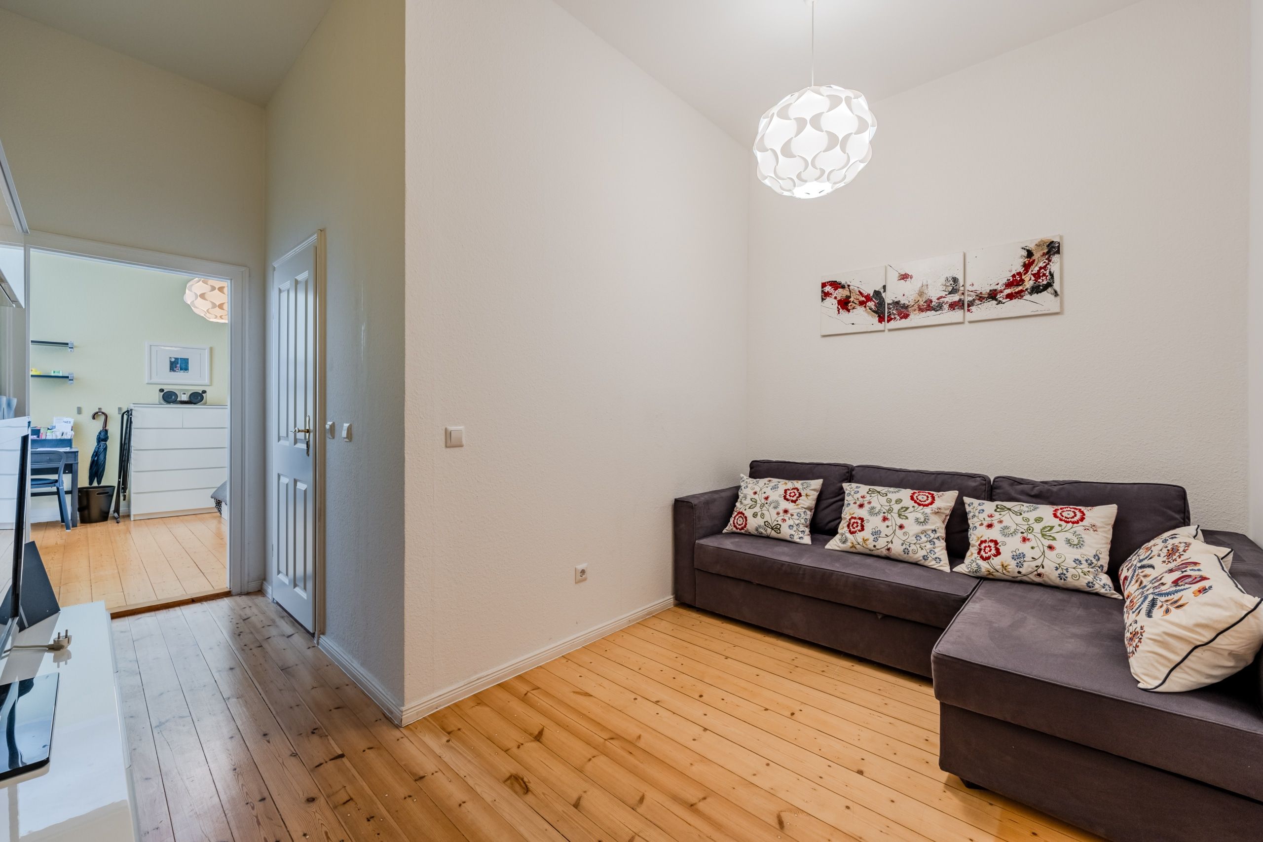 Wohnzimmer aus Eckperspektive mit Holzdielenboden und grauem Ecksofa sowie dreiteiligem Wandbild