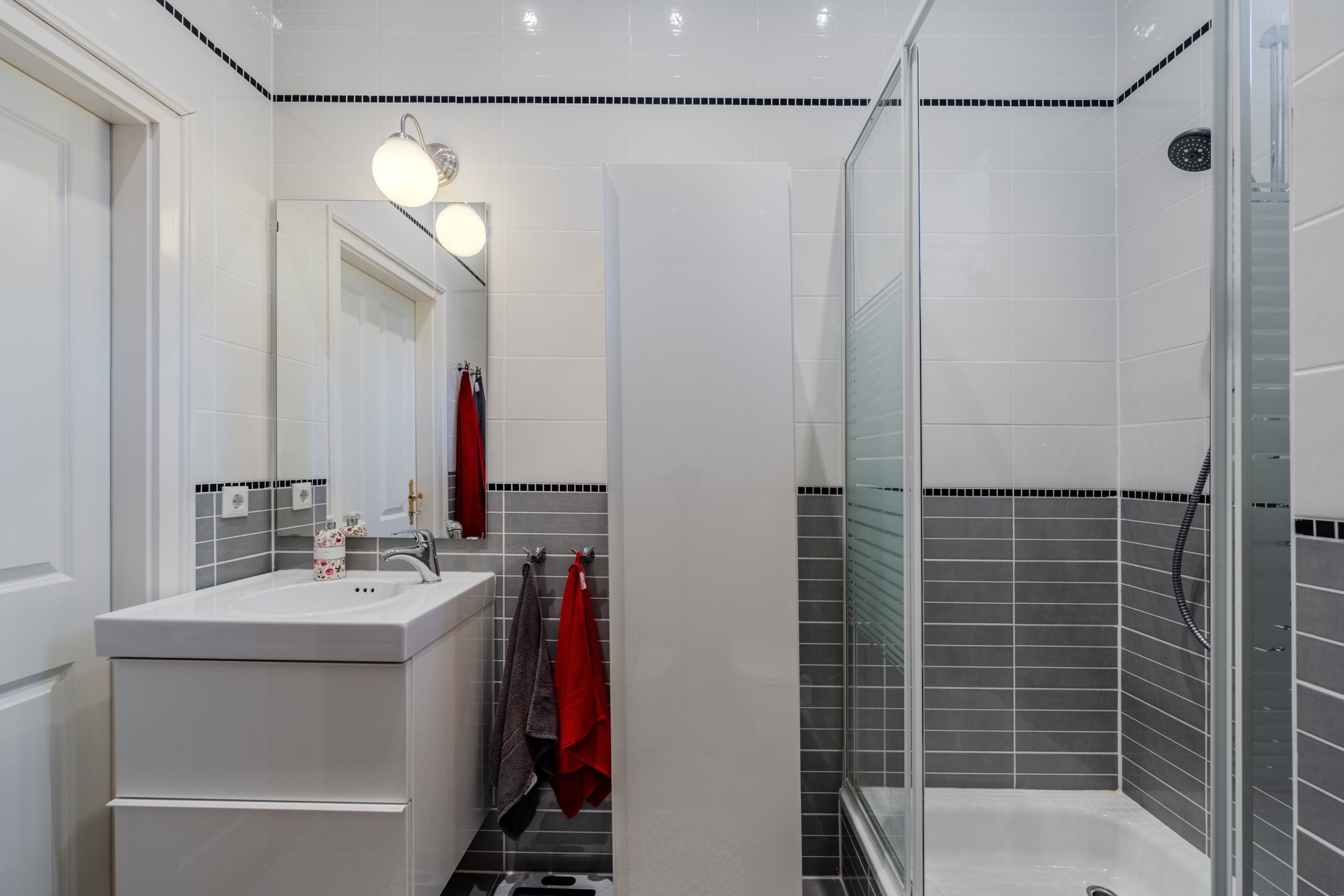 Innenansicht Badezimmer mit Glas-Duschkabine weißem Waschbeckenunterschrank grauen Wandfliesen und Handtuchhaken