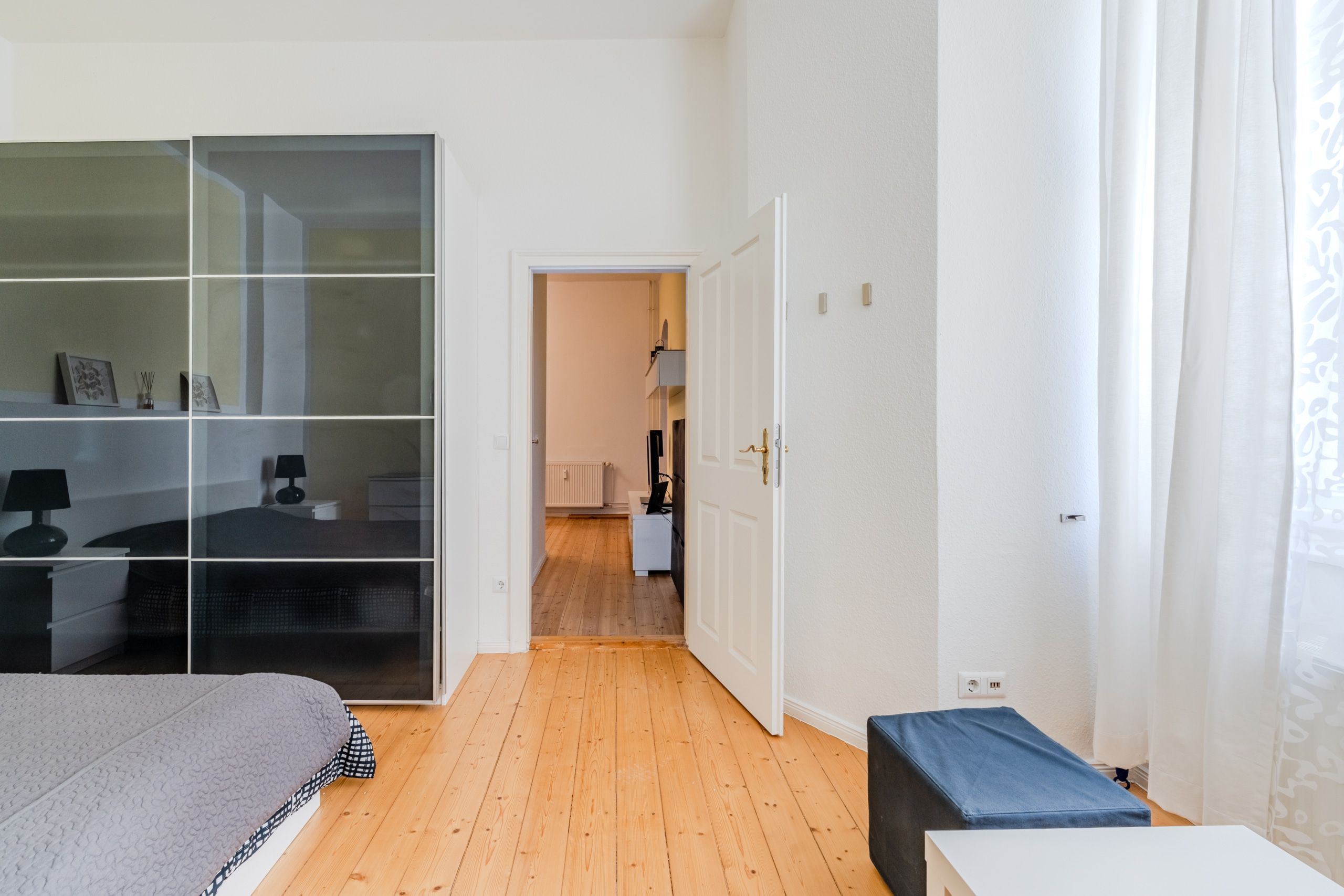 Innenaufnahme eines Schlafzimmers mit Holzdielen, schwarzem Spiegelschrank, offenen weißen Türen zum Wohnzimmer