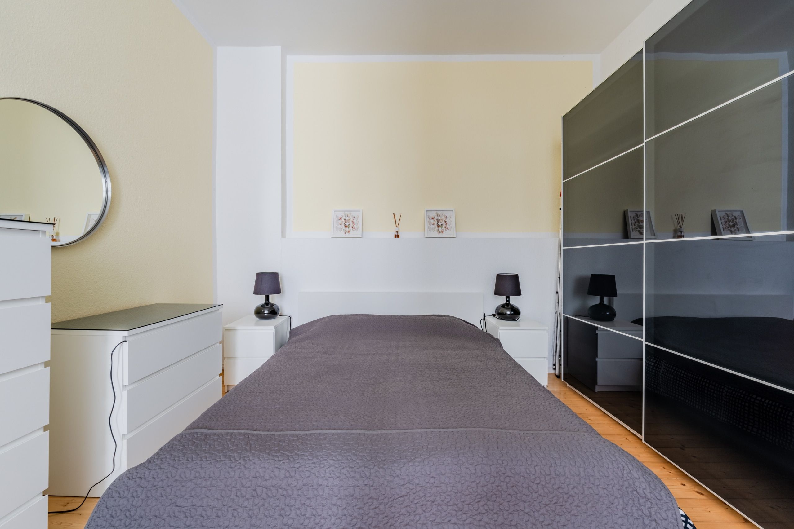 Schlafzimmer frontal mit grauem Einzelbett, rundem Spiegel, weißem Nachttisch und schwarzem Schiebetürenschrank