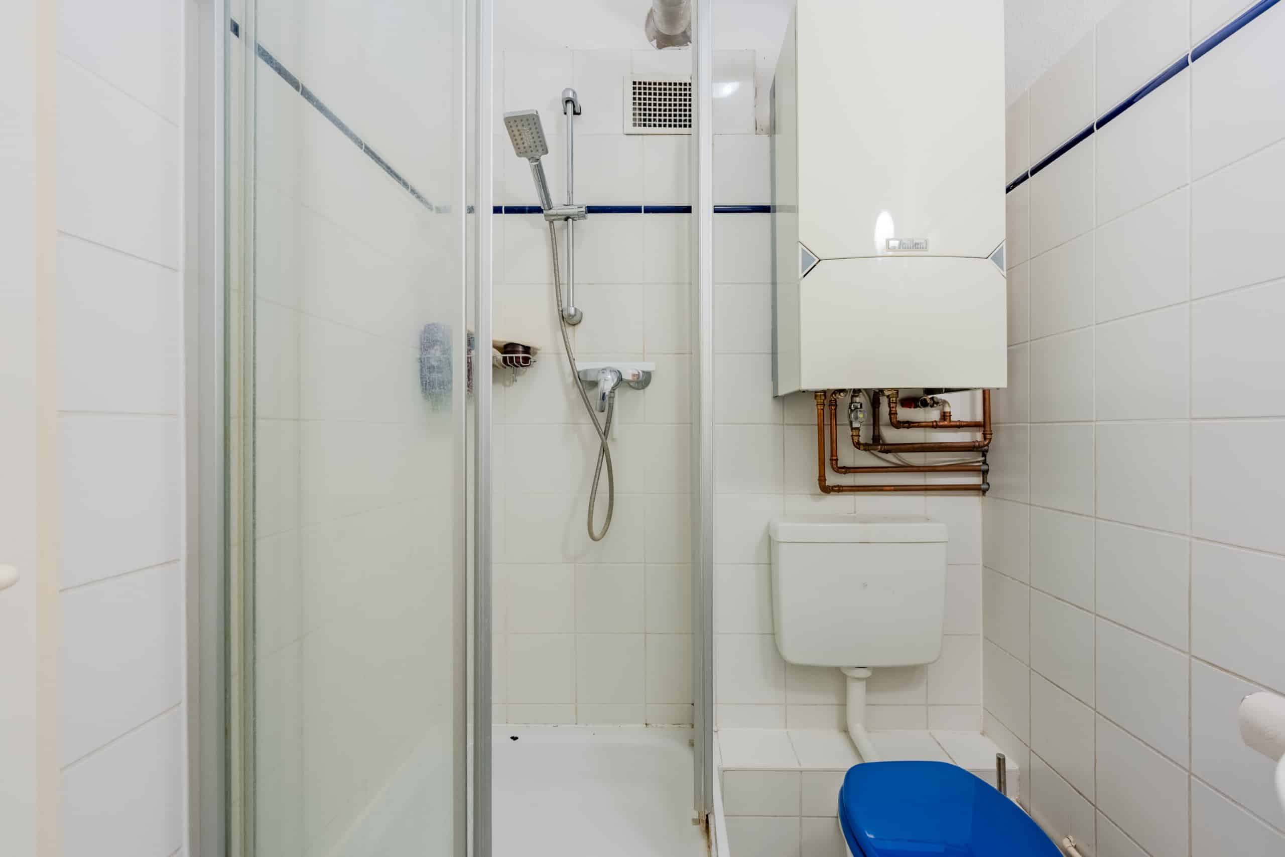 Innenansicht eines Badezimmers mit Glastürdusche wandhängendem Boiler und WC mit blauem Sitz