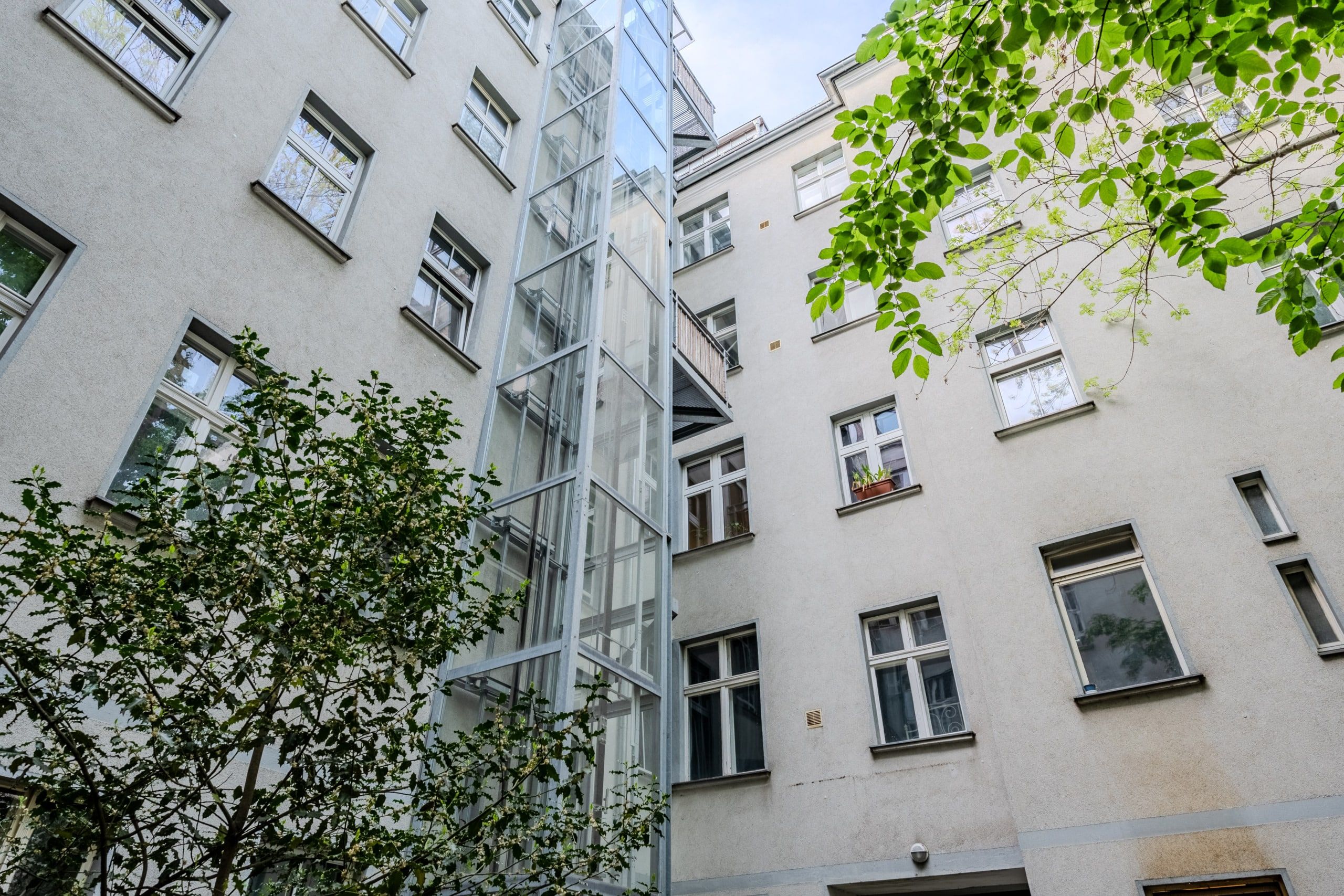 Innenhofansicht mehrgeschossiger Wohnhausfassade mit grauer Putzfassade und freistehendem Glasaufzugsturm