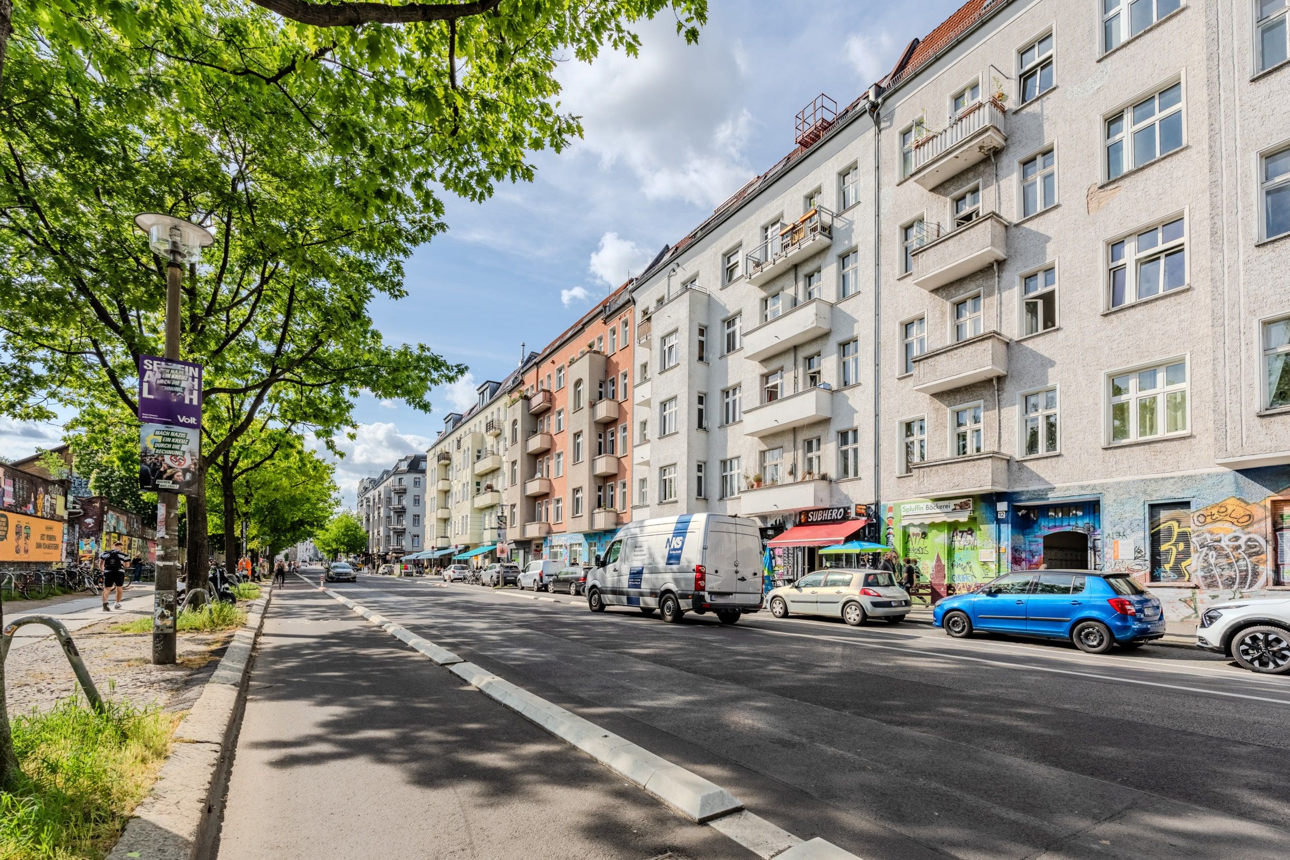 Straßenansicht von mehrgeschossigen Wohnhäusern mit Balkonen und Erdgeschossladen unter begrünten Bäumen