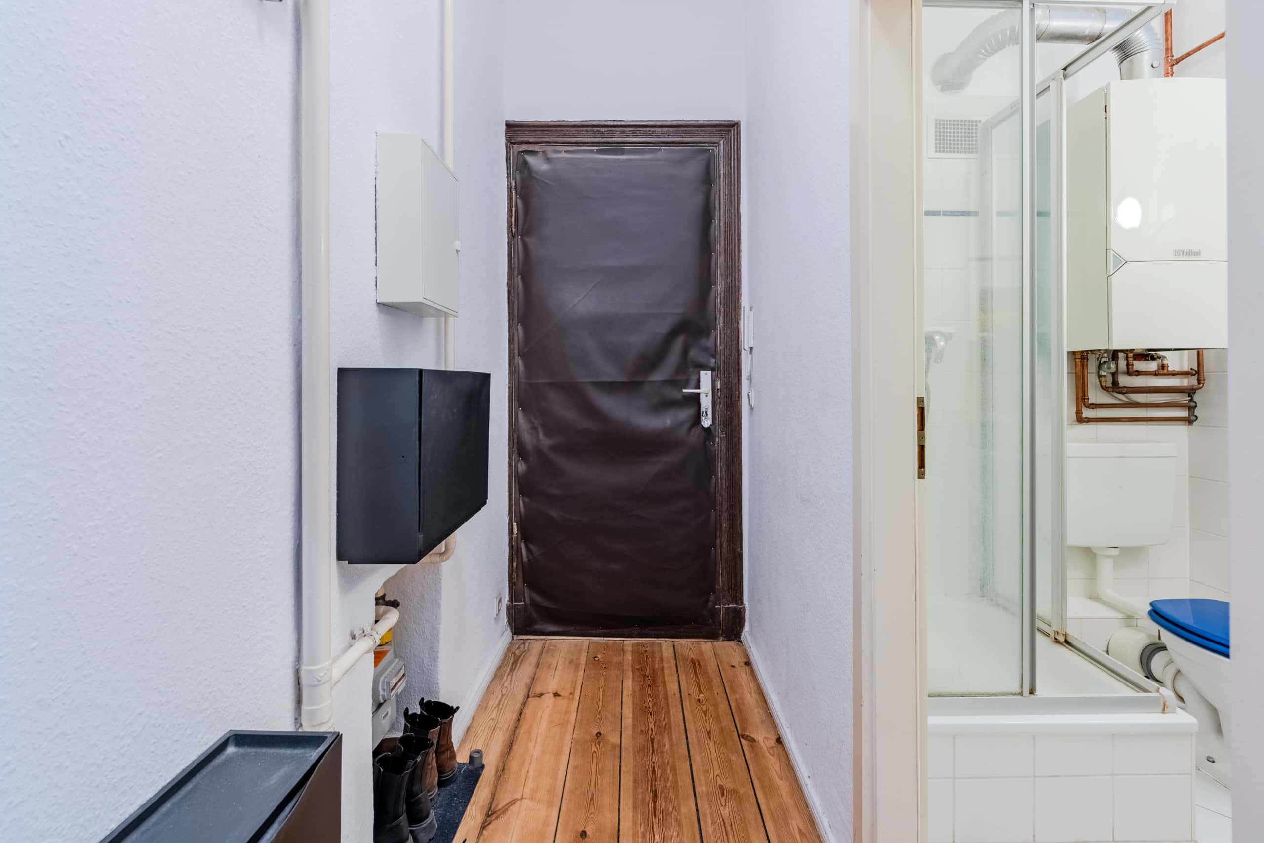 Innenflur einer Altbauwohnung mit brauner Eingangstür, offener Duschkabine, WC und sichtbarer Heiztherme
