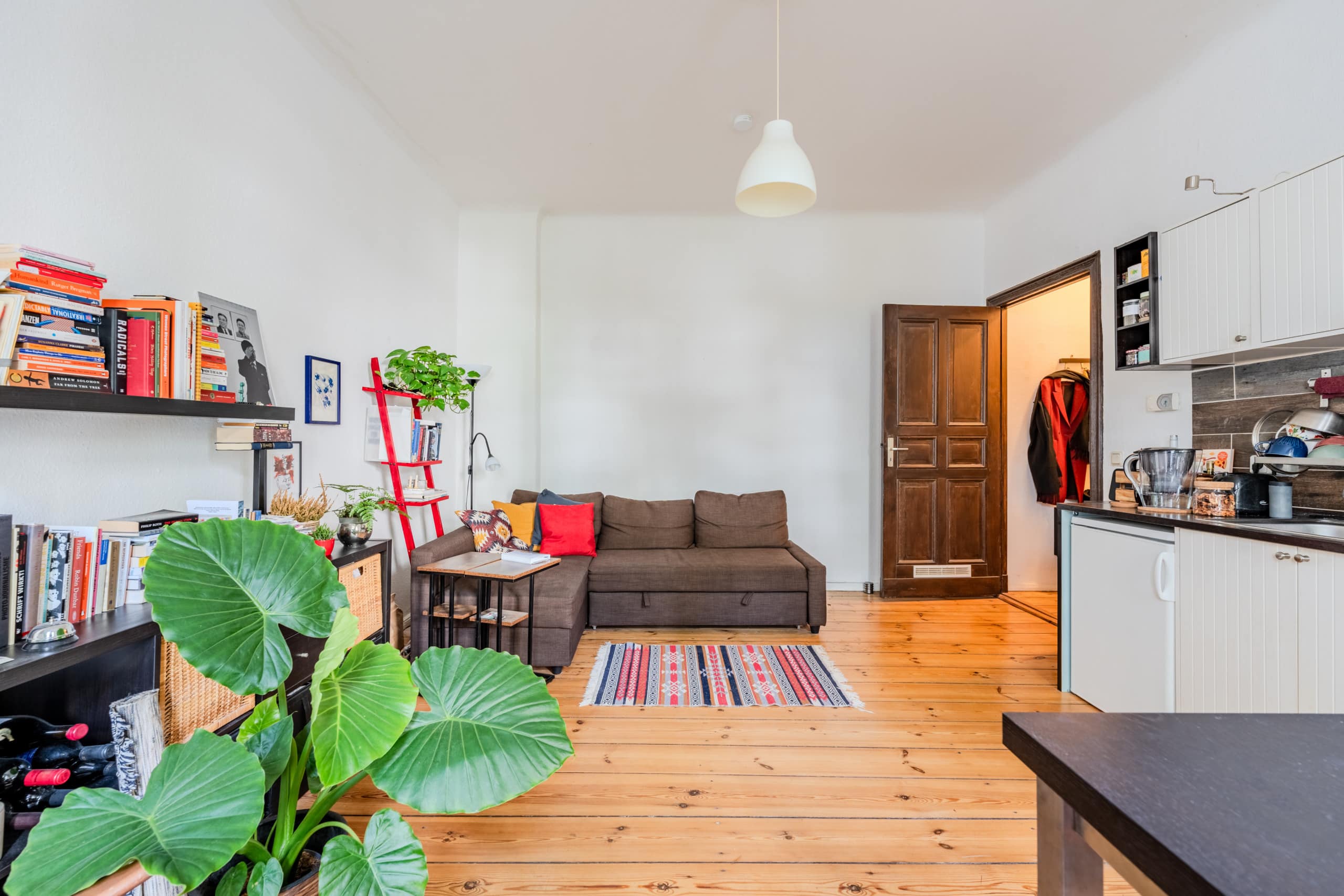 Heller Wohnraum einer Altbauwohnung mit Ecksofa, offener Küchenzeile, Bücherregal und Parkettboden in Frontalansicht