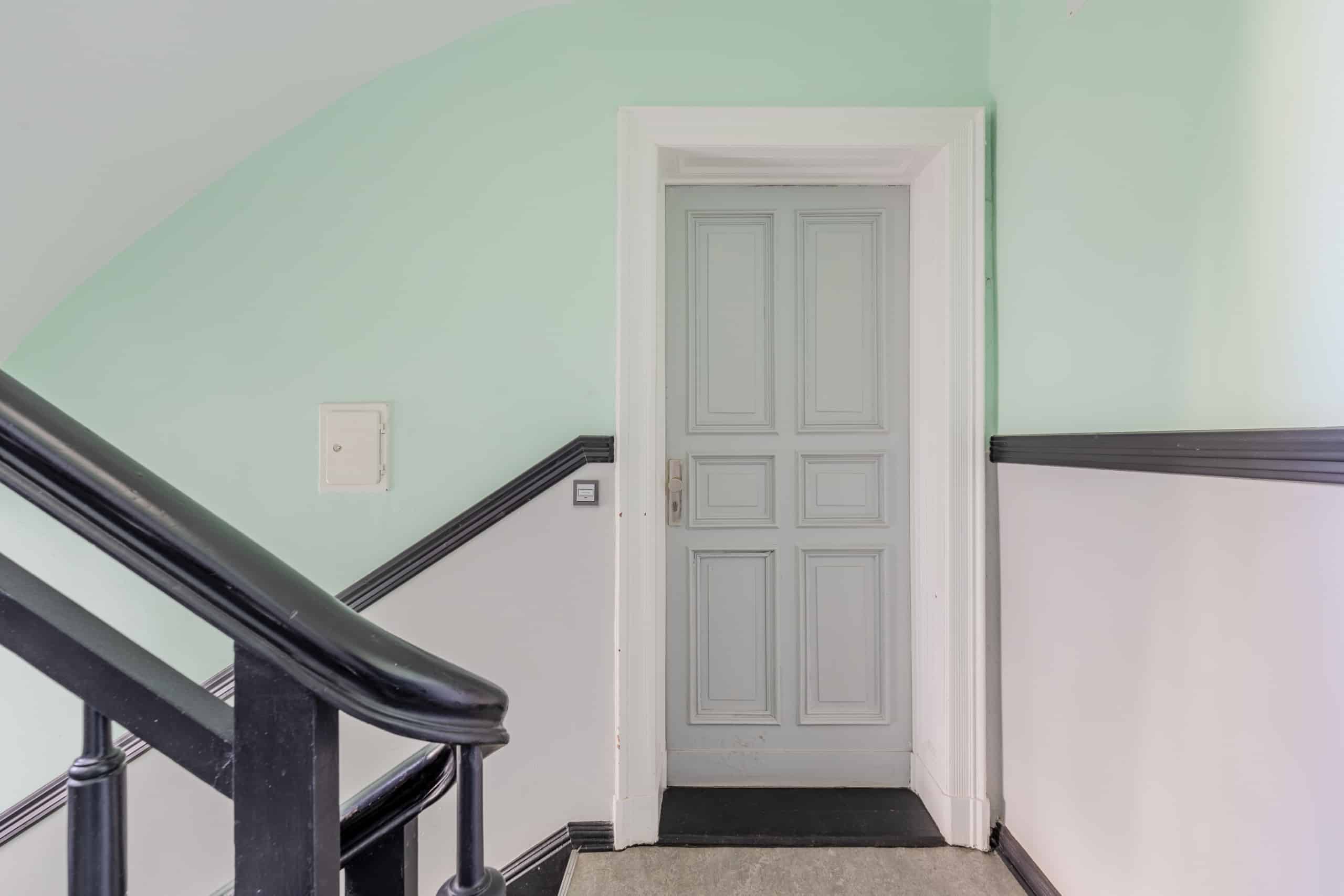 Blick auf historische Paneeltür im Altbauflur mit pastellgrüner Wand und schwarzem Holzgeländer