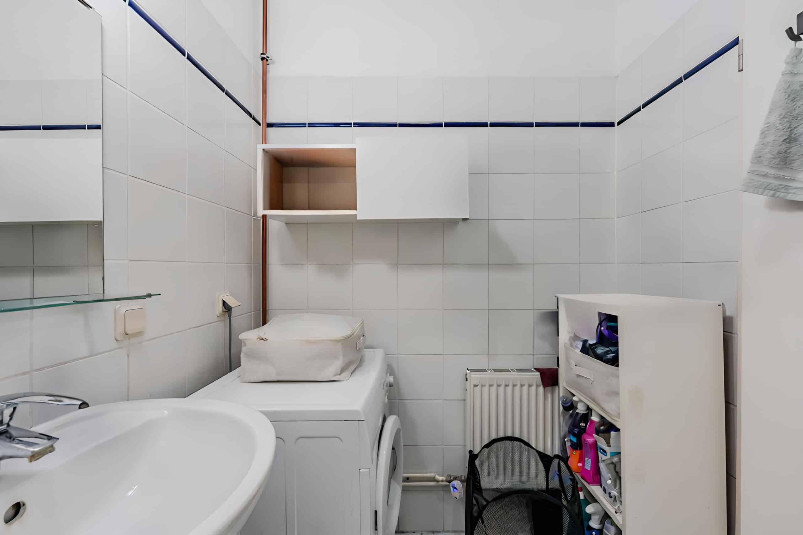 Frontale Ansicht kleines Badezimmer mit Waschmaschine weiße Wandfliesen blauer Bordüre offenes Wandregal und Wäschekorb