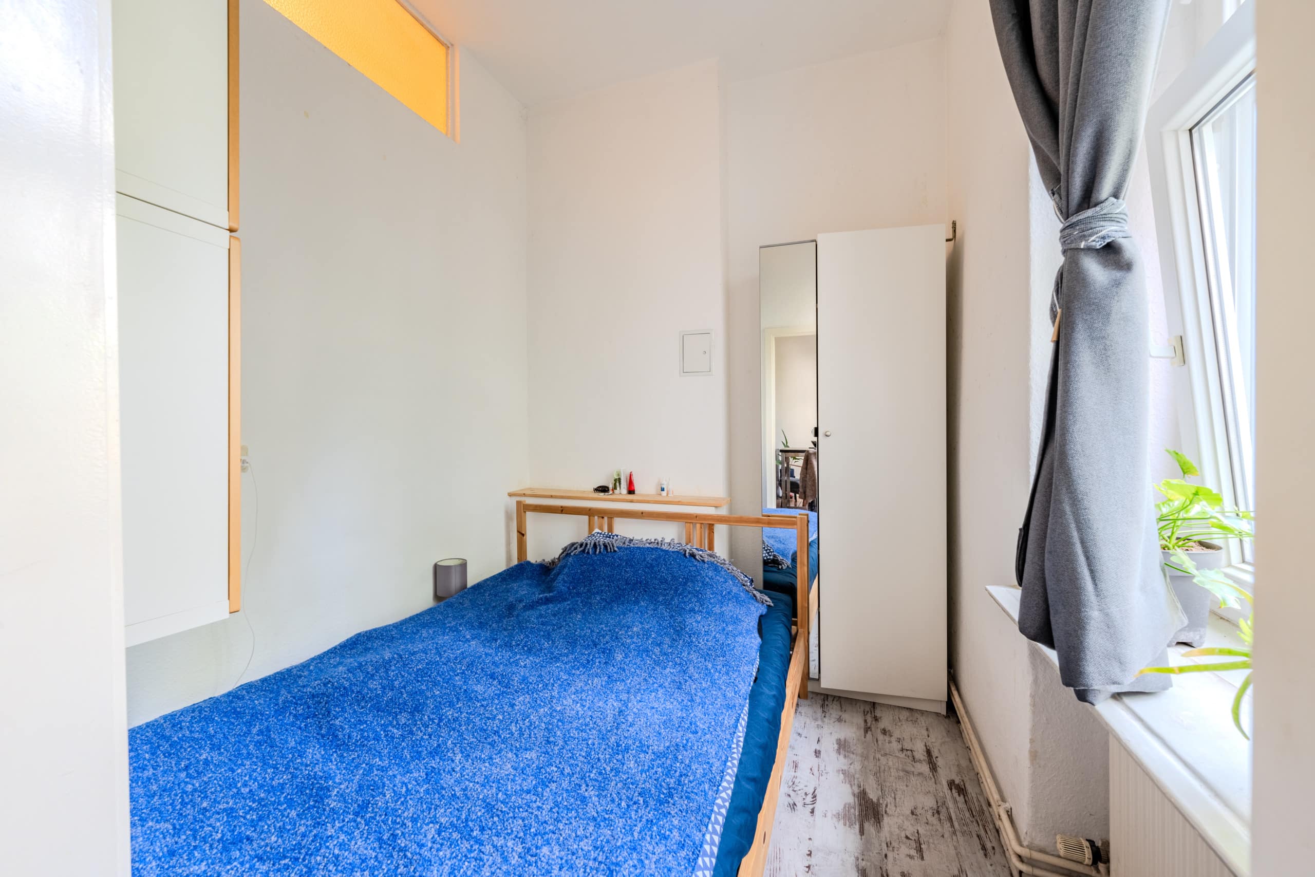 Innenraum eines kleinen Schlafzimmers mit Holzbett, blauem Überwurf, weißem Spiegelschrank und Vorhangfenster