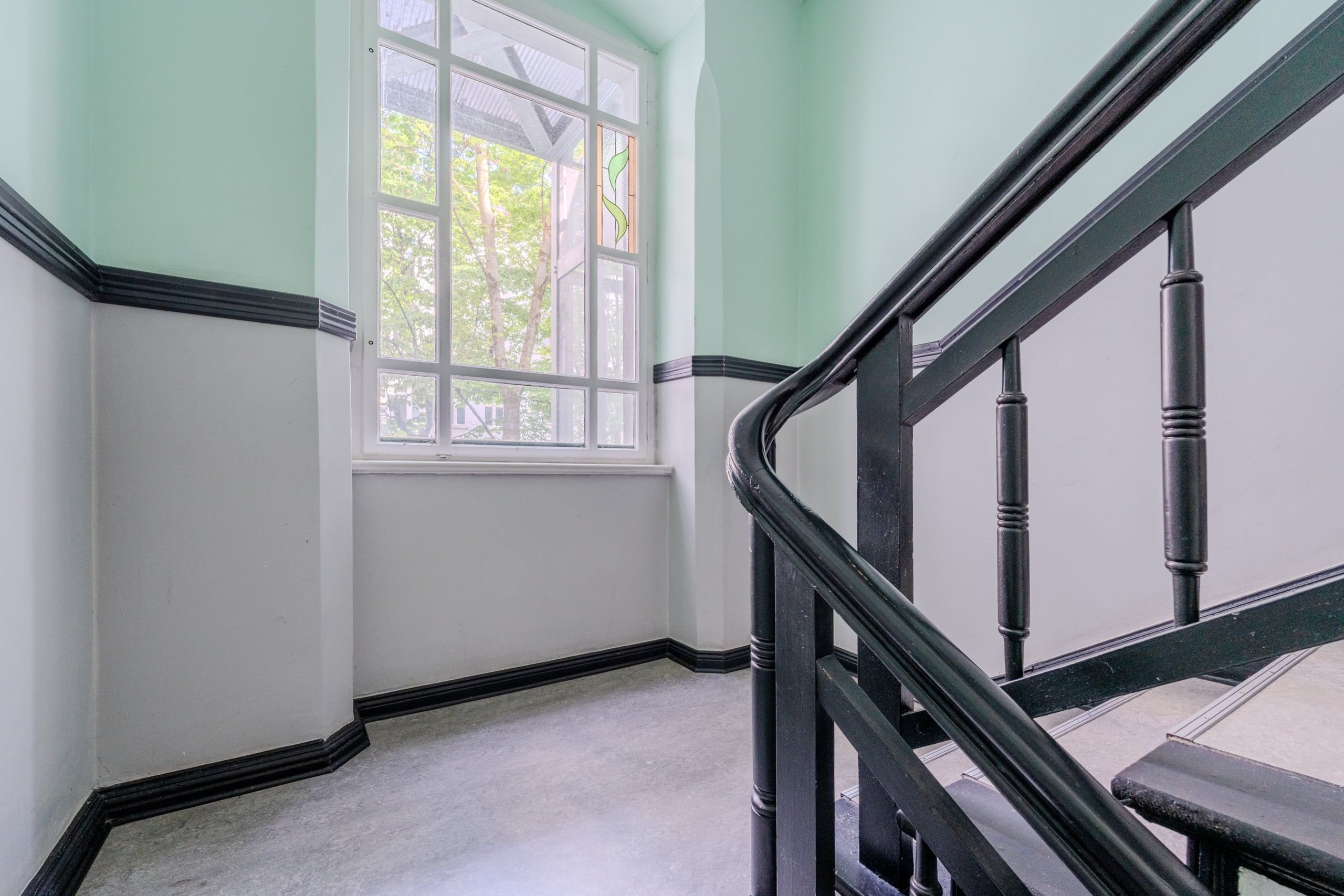 Treppenhaus mit geschwungenem schwarzem Holzgeländer und großem gläsernen Sprossenfenster mit grünem Motiv