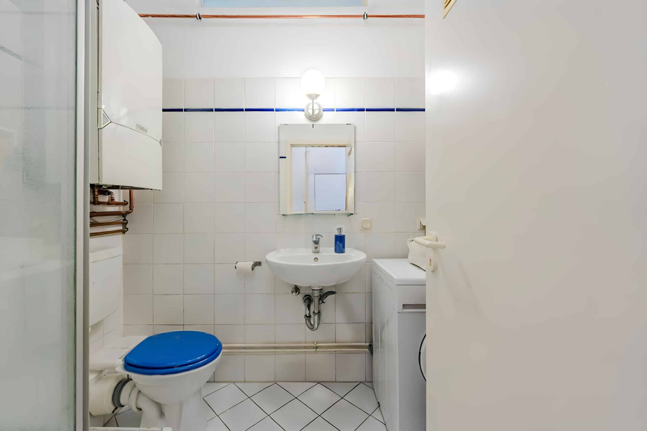Kompaktes Badezimmer mit weißen Fliesen, blauem Toilettensitz, Waschbecken, Spiegel und Waschmaschine in Frontansicht