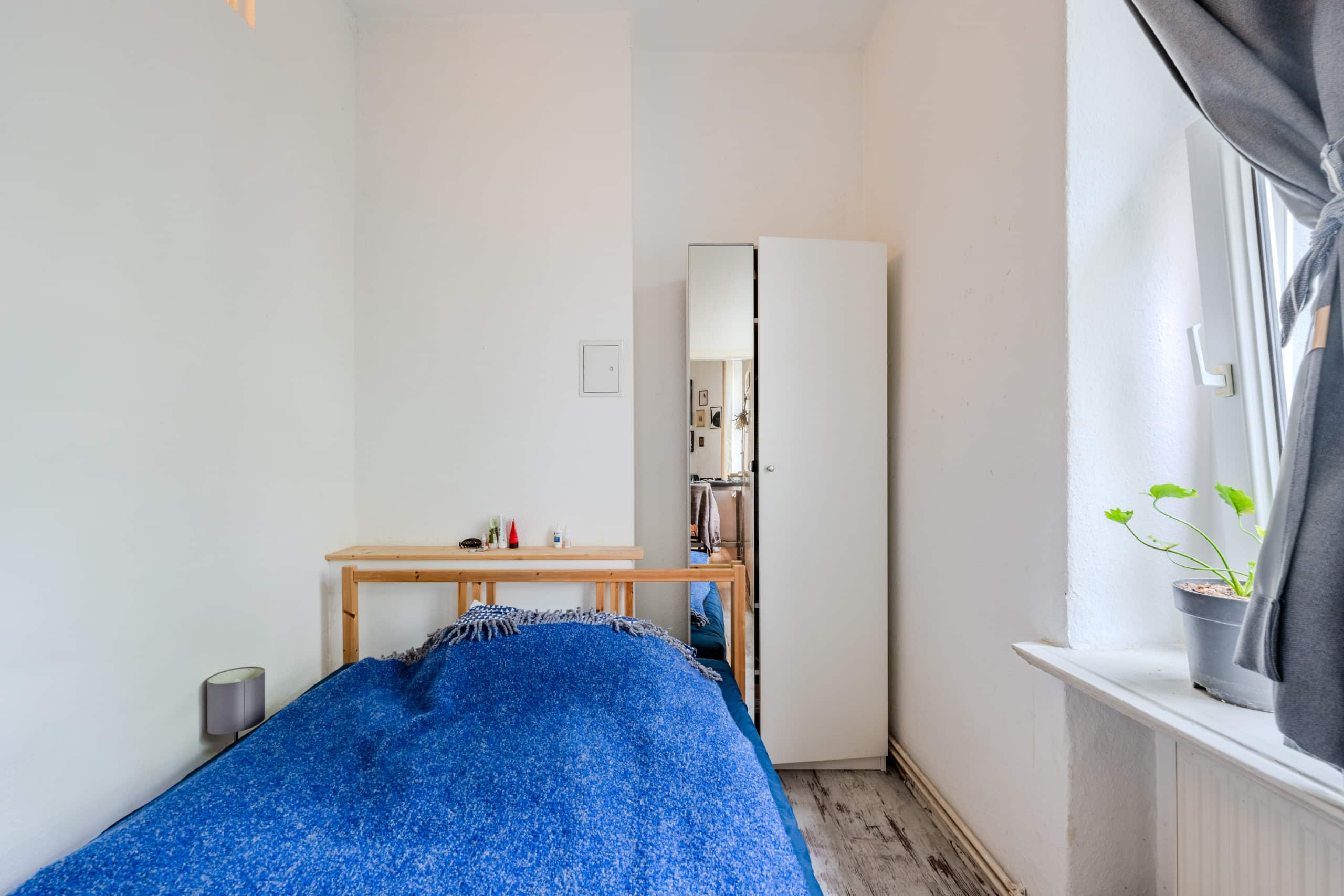 Schlafzimmer mit Einzelbett (Holzrahmen), blauer Decke, weißem Spiegelschrank und Fenster mit Topfpflanze