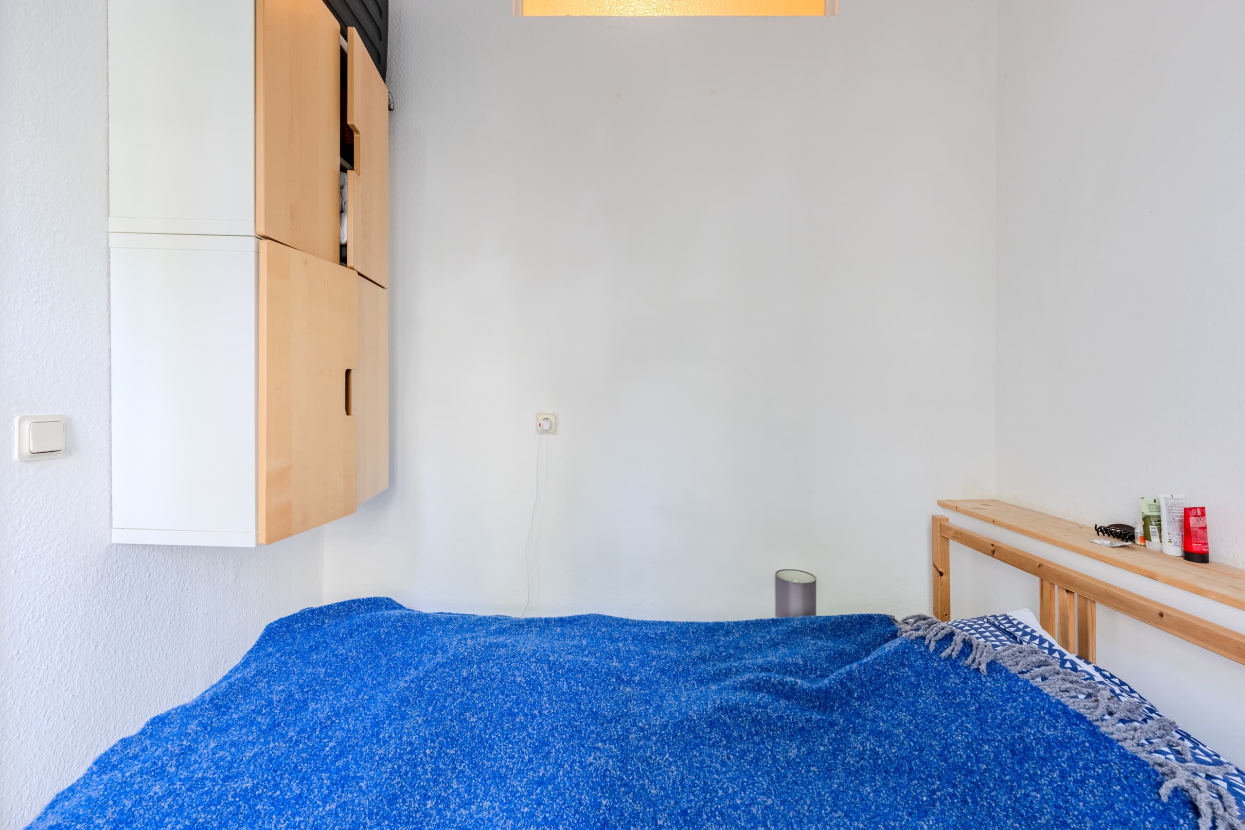 Innenansicht Einzelzimmer mit blauem Bettüberwurf wandhängendem Holz-Weiß-Schrank und Holzregal als Kopfteil