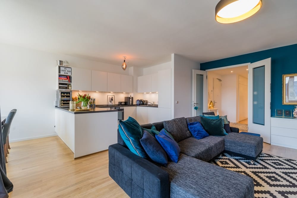 Blick in offene Wohnküche mit Kücheninsel, grauem Ecksofa, Sideboard vor türkisfarbener Akzentwand und Parkettboden