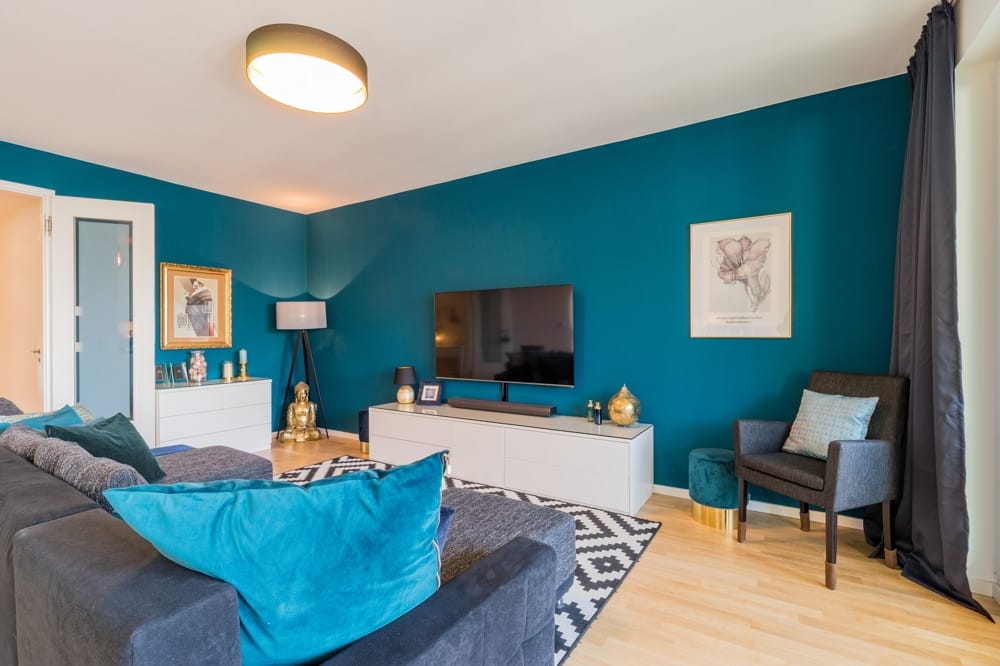 Modernes Wohnzimmer einer Mietwohnung frontal mit petrolfarbener Wand, grauem Sofa, TV-Möbel und Sessel