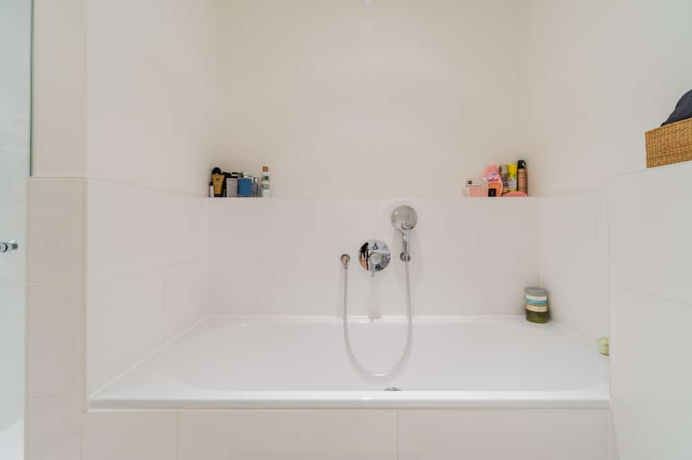 Badezimmer mit eingelassener Badewanne, verchromter Handbrause und seitlichen Nischen mit Pflegeprodukten
