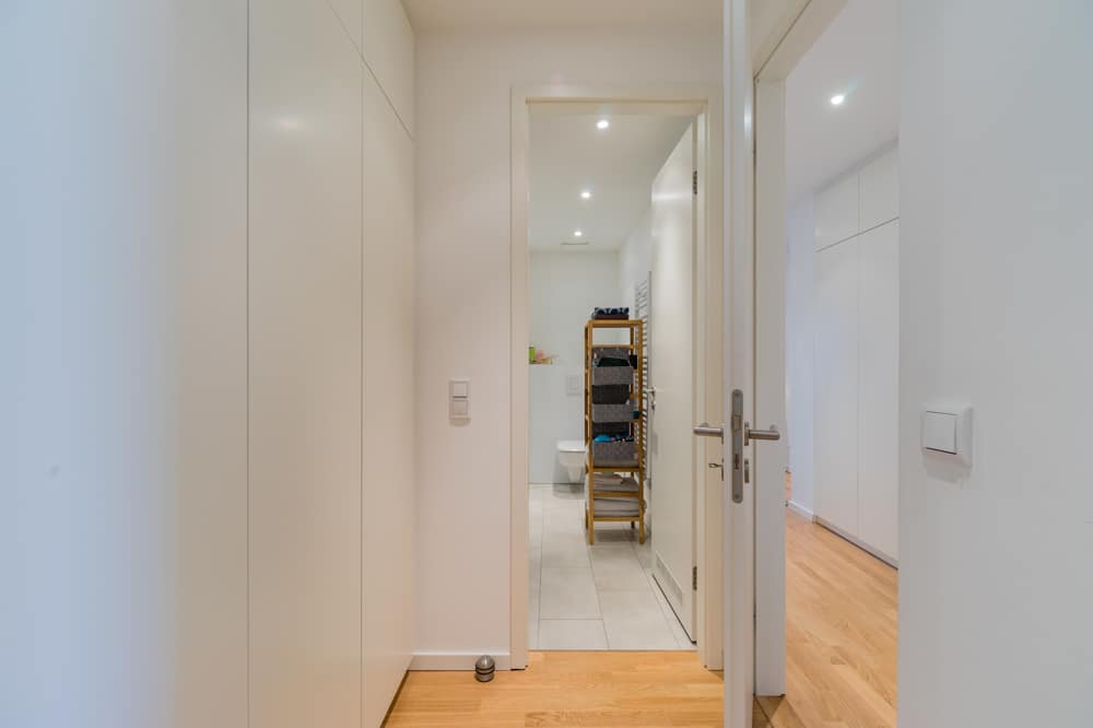 Innenansicht einer Wohnung mit Blick vom Flur in ein Bad weiße Einbauschränke Holzboden und offenes Regal